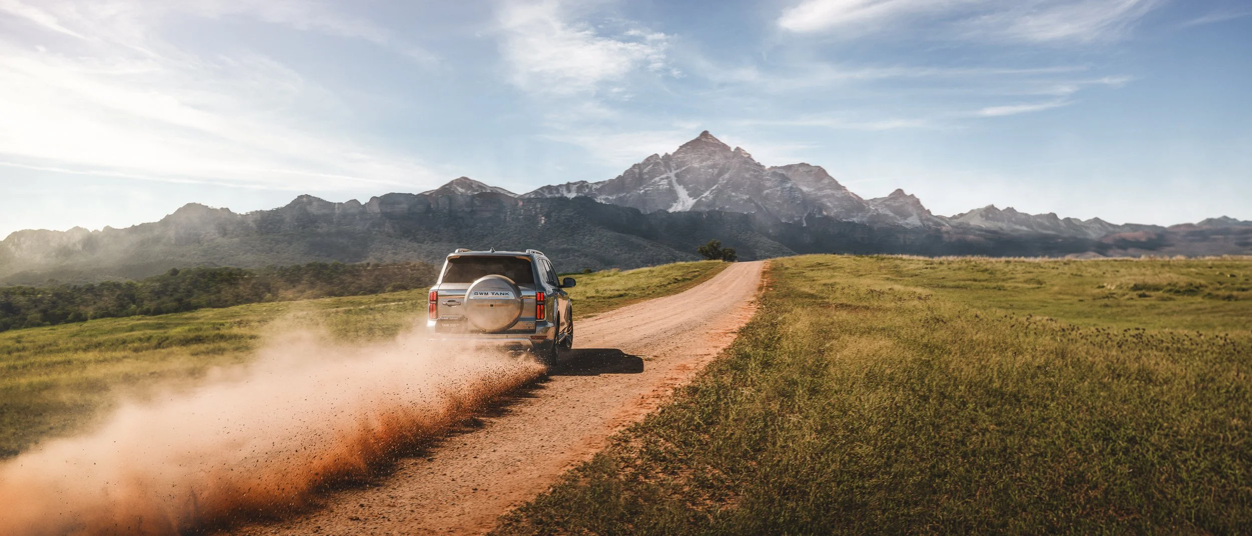 An SUV driving on a dirt road in a grassy field with mountains in the background under a partly cloudy sky.