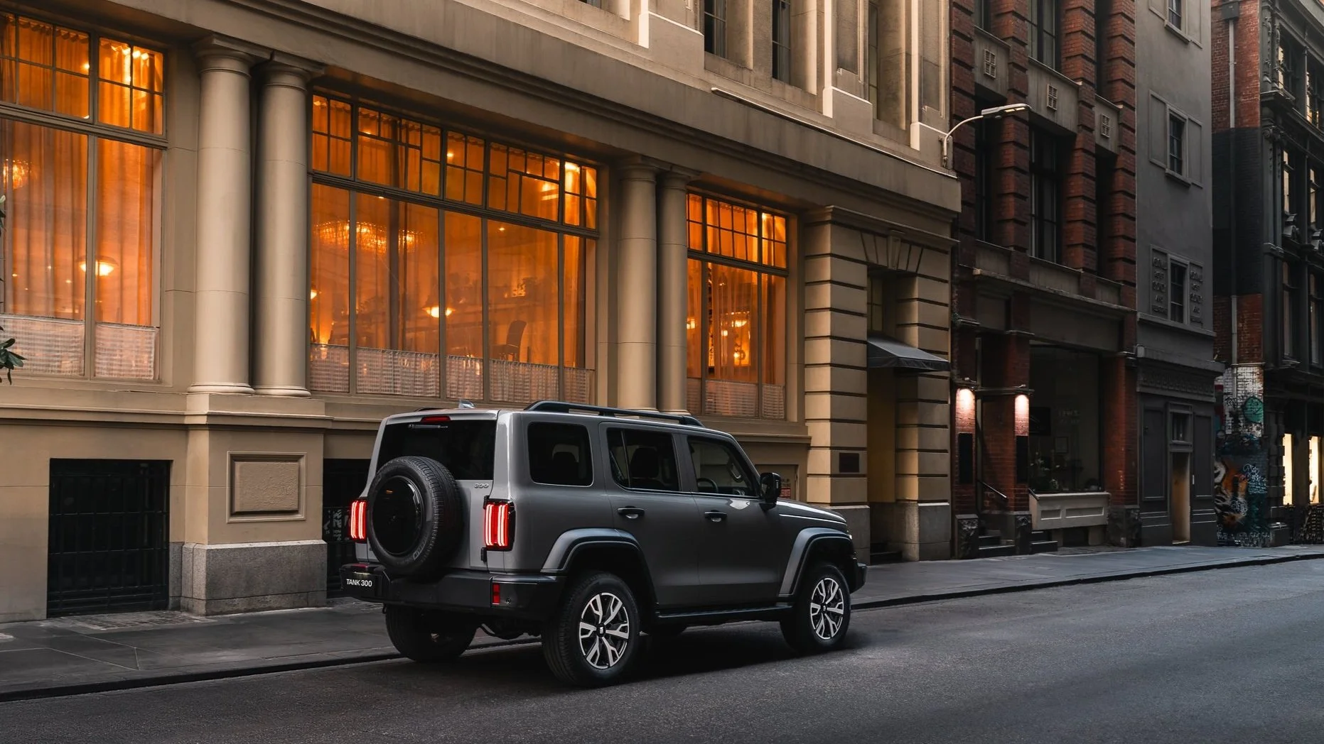 Gray Jeep SUV parked on city street in front of illuminated building with large orange windows and classical columns.