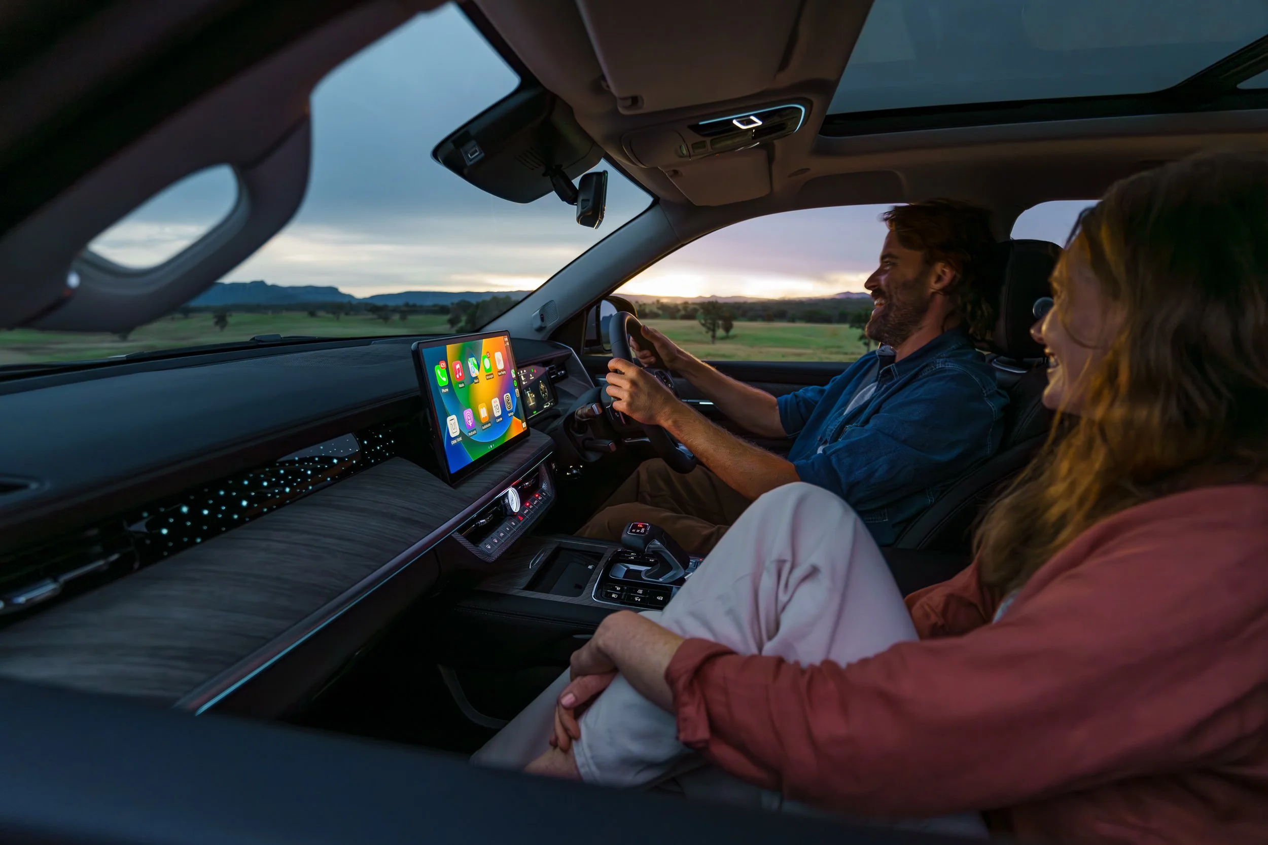 Interior of a car with two passengers, a man at the driver’s seat and a woman in the passenger seat, during sunset with a scenic landscape outside. The dashboard features a large touchscreen with app icons.