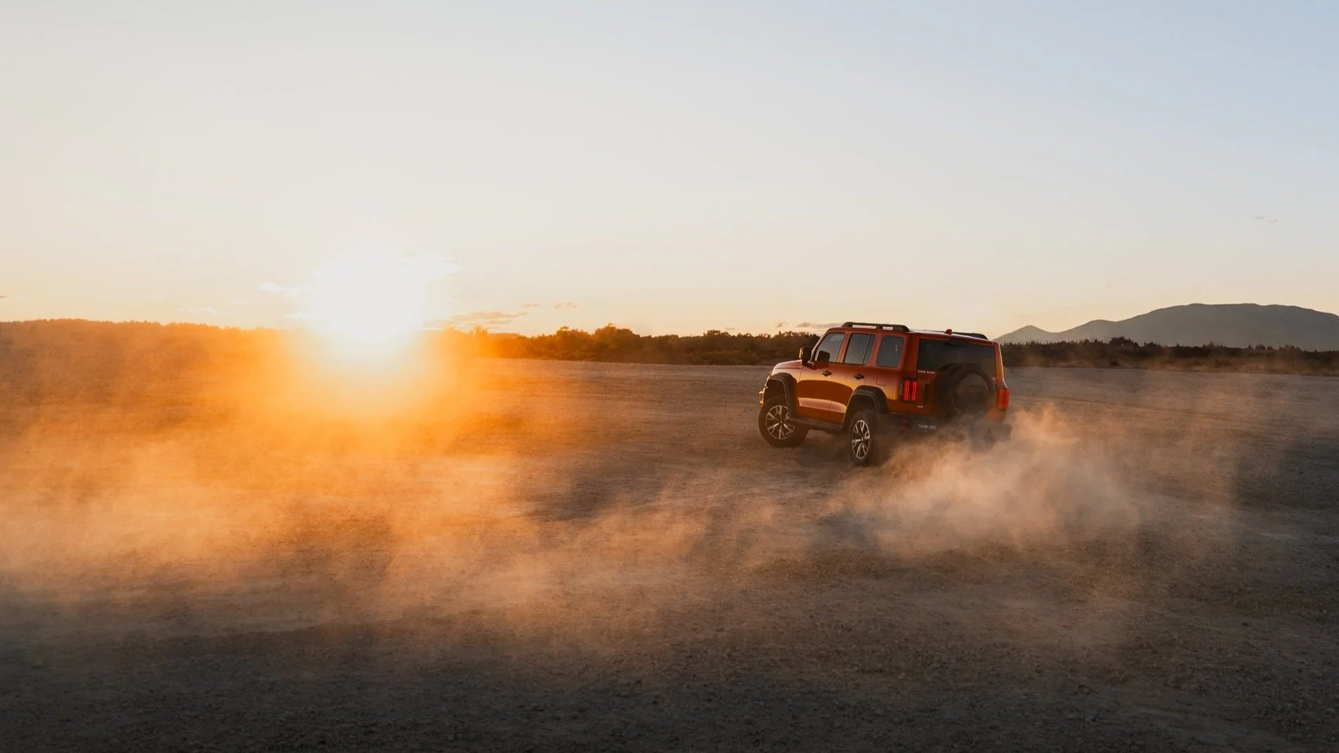 An orange SUV driving across a dusty, open field at sunset with mountains in the background.