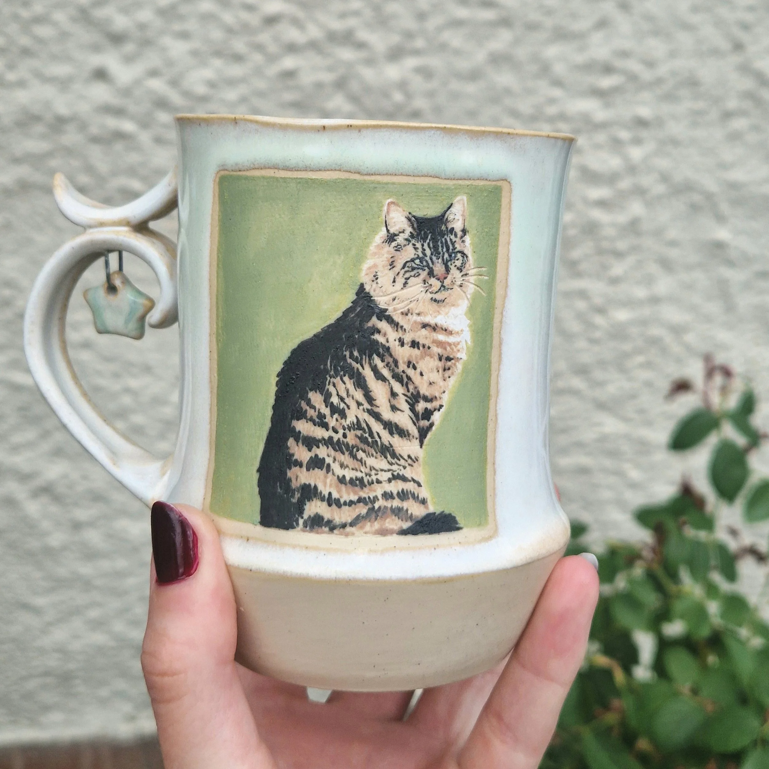 Close-up of a ceramic mug decorated with a painted image of a tabby cat, held by a person with painted nails, against a blurred outdoor background.