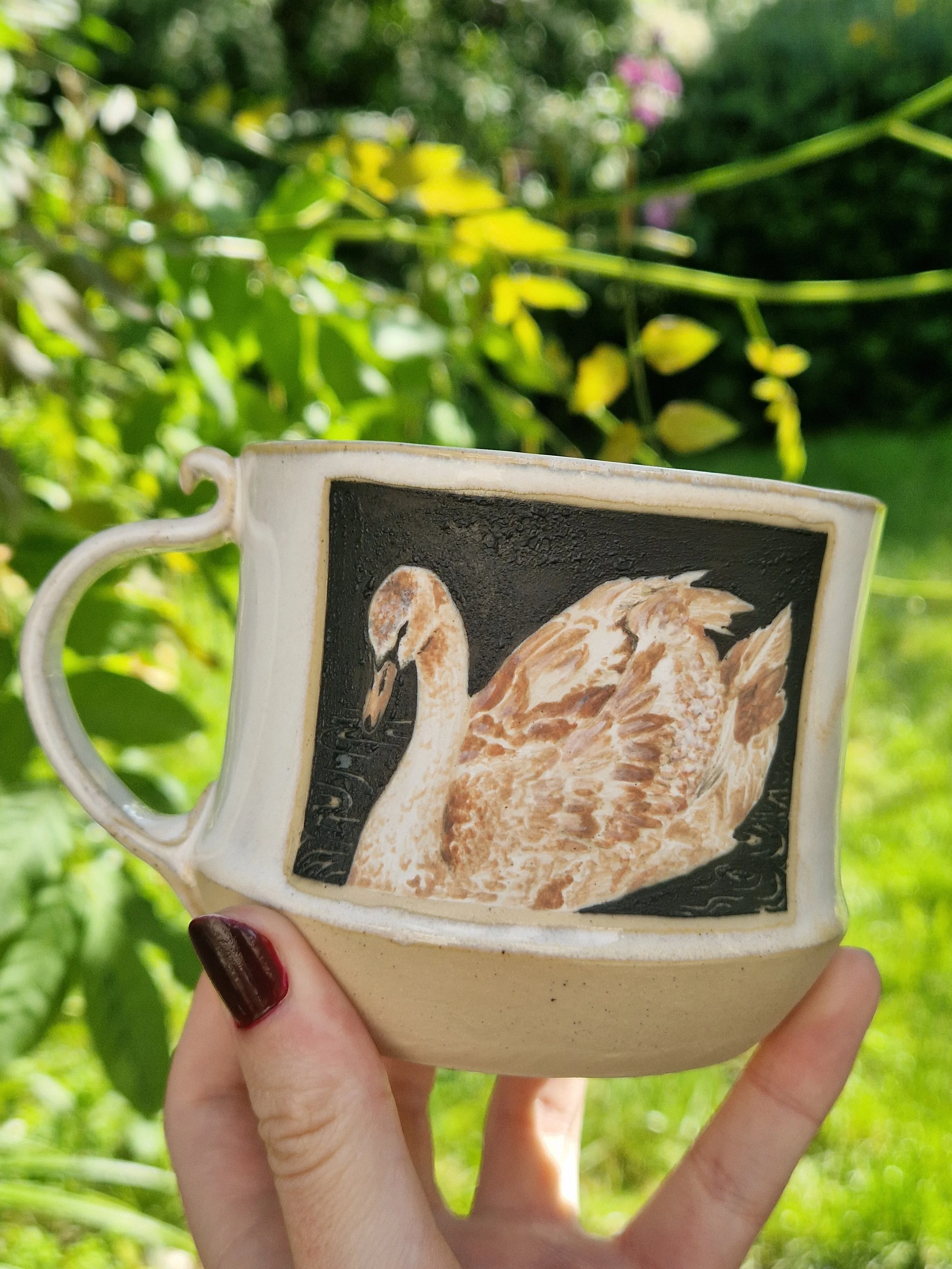 A person with dark red painted nails holding a ceramic mug with a coffee bean illustration. The mug has a cream-colored handle and is being held outdoors against a background of green foliage and some purple flowers, with sunlight filtering through.