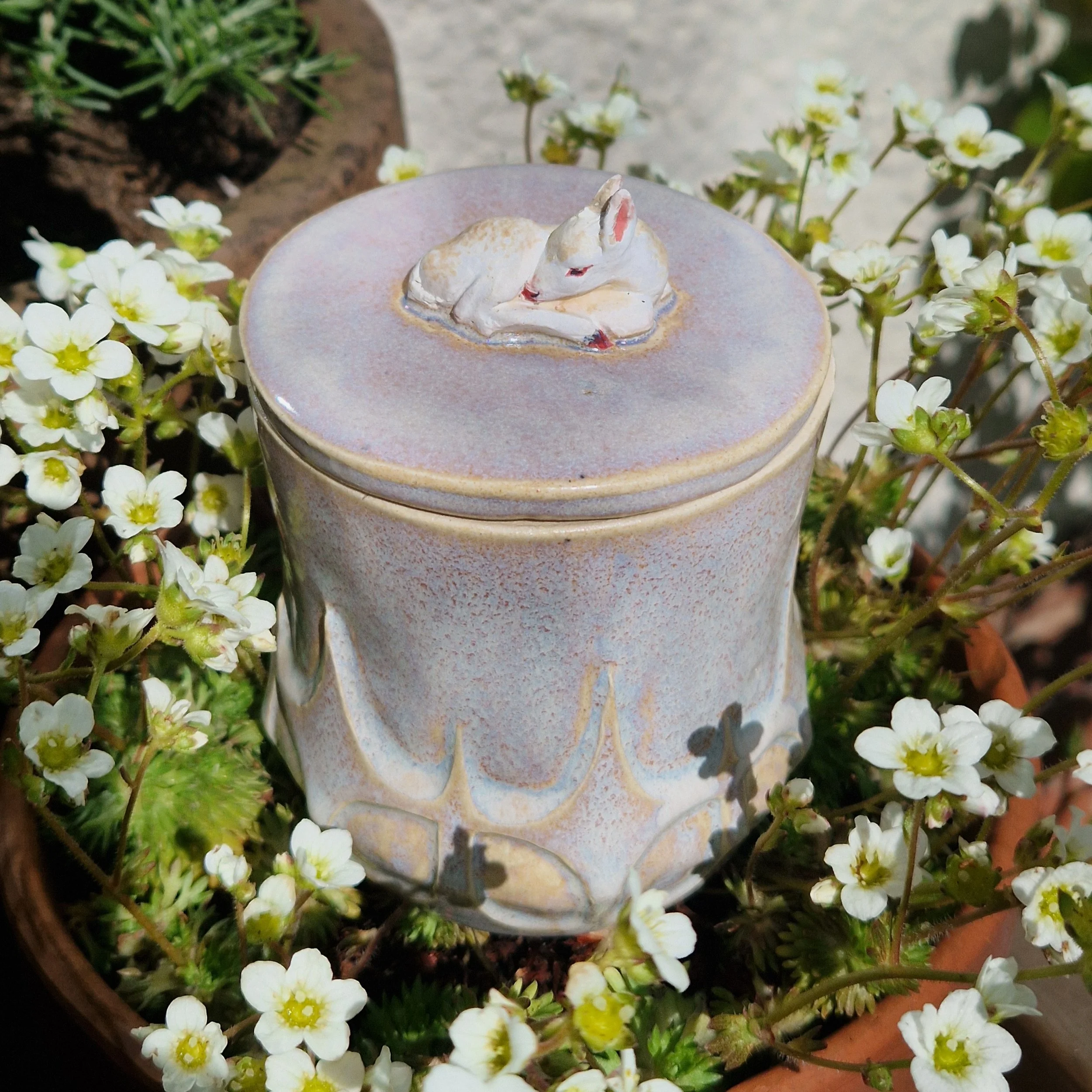 A small ceramic jar topped with a sculpture of a sleeping white fawn, surrounded by green plants and white flowers in a terracotta pot.