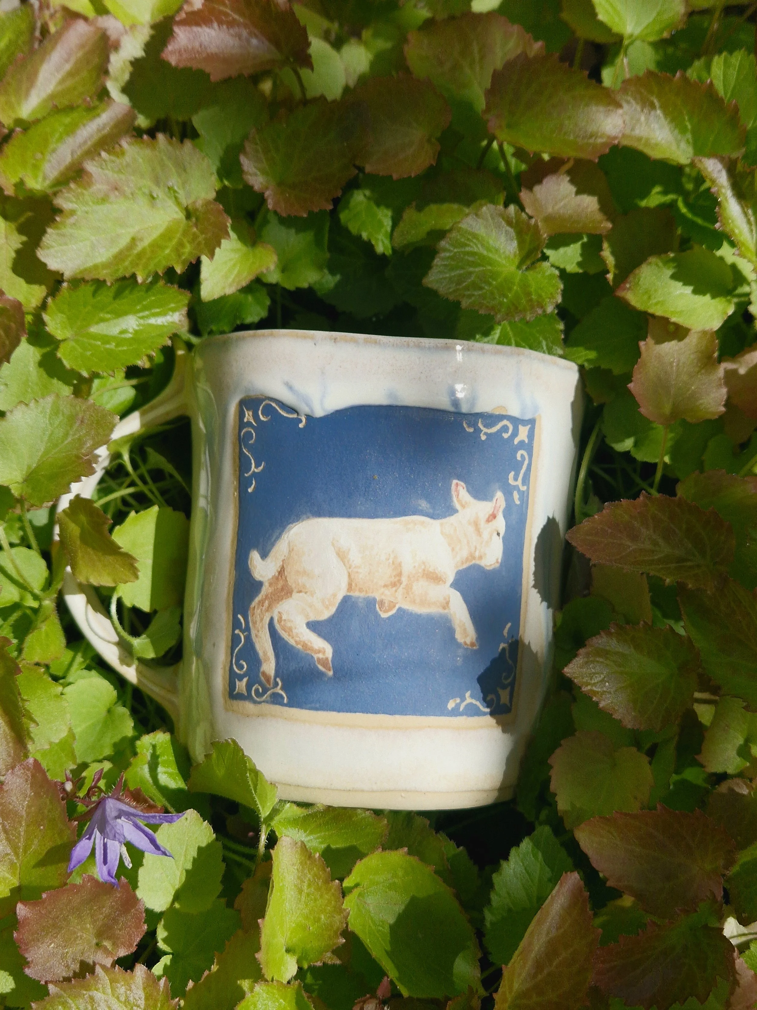 A white ceramic mug with a picture of a frolicking lamb on a sky blue background, surrounded by green and reddish leaves.
