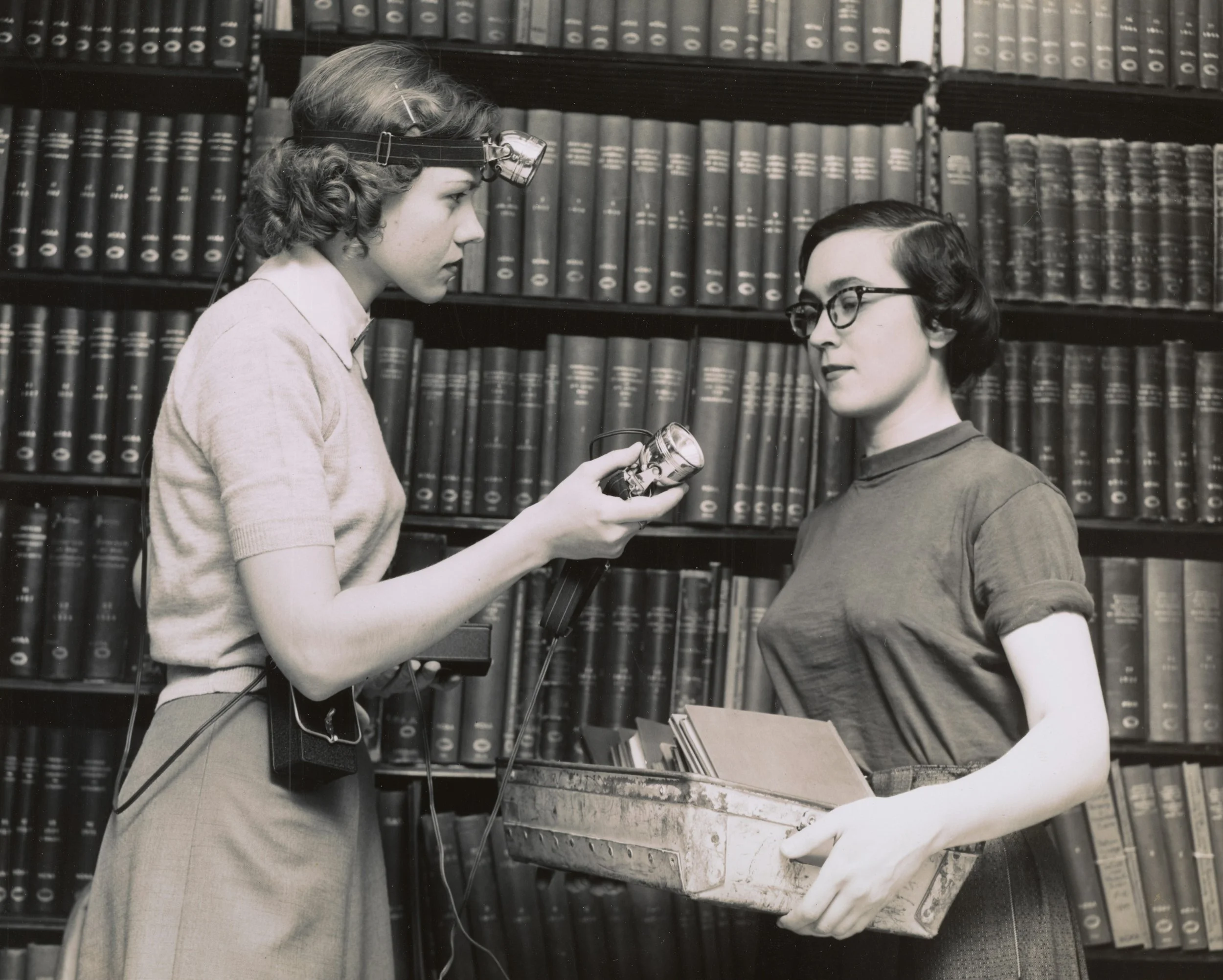 Two women, one with curly hair and glasses, the other with short wavy hair and a headlamp, standing in a library with shelves of books, inspecting a book