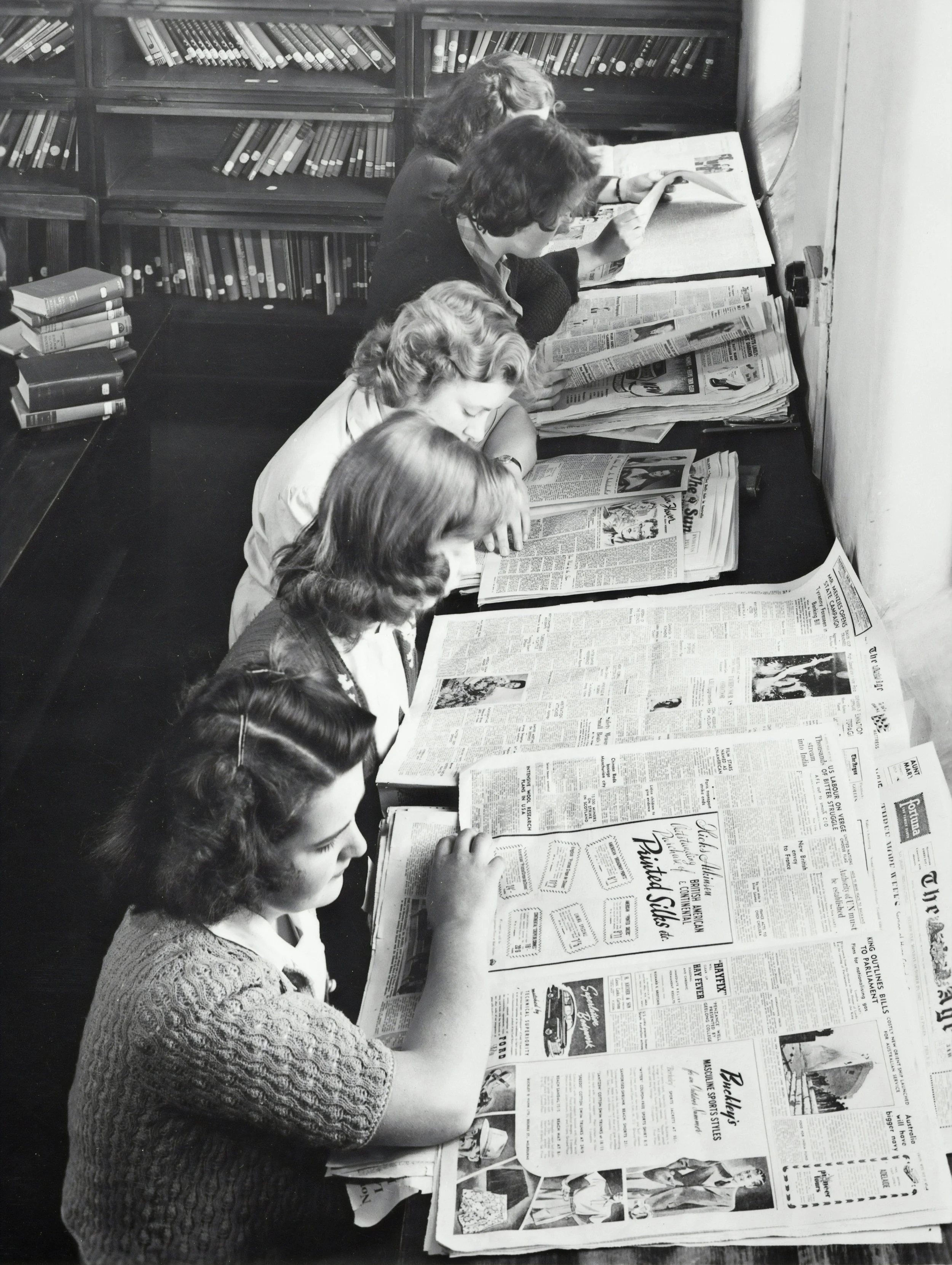 A black-and-white photo shows six women seated at a long table in a library or study area, reading newspapers. The table holds numerous newspapers and some books. Behind them are shelves filled with books.