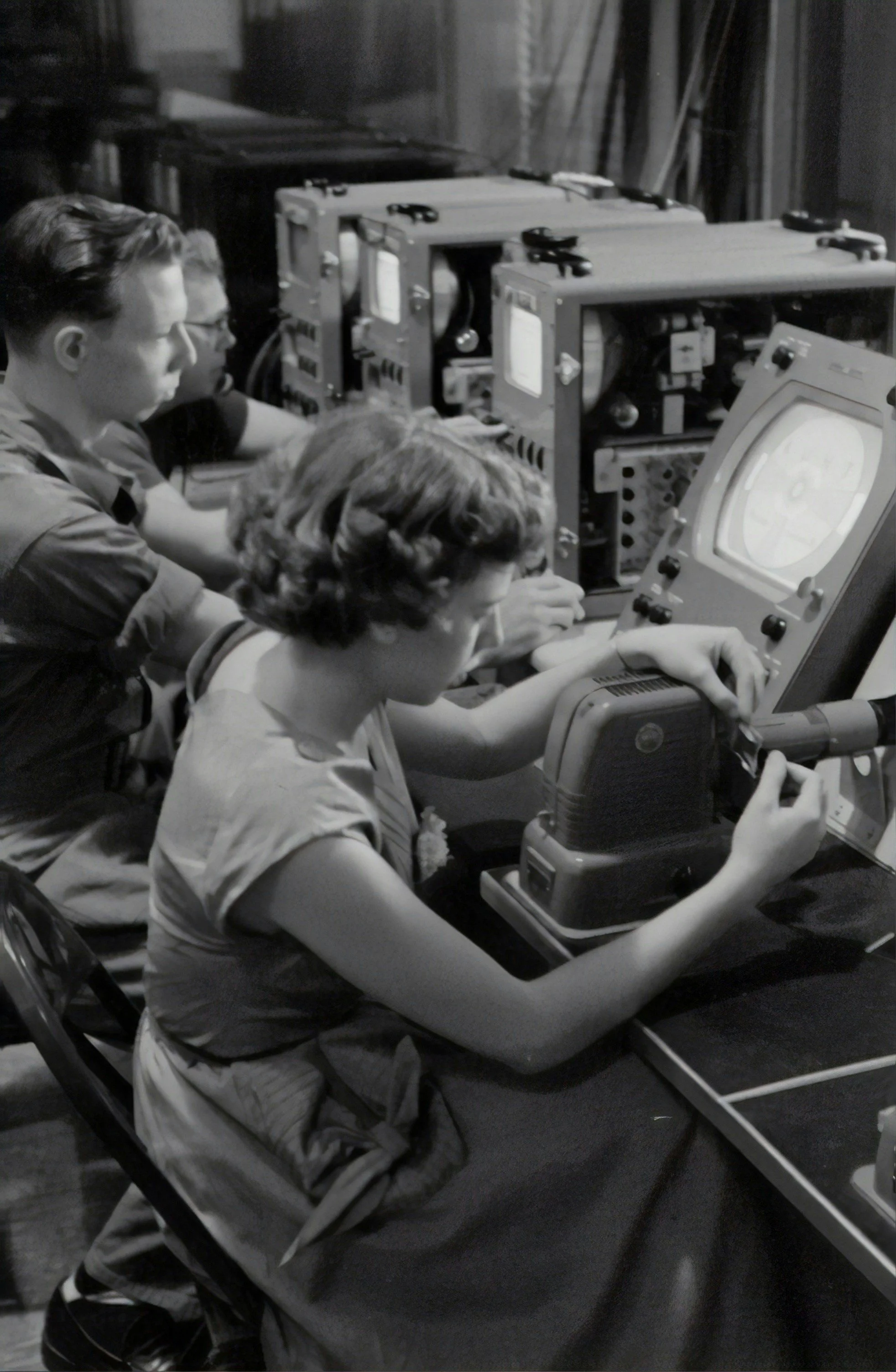 Black and white photo of three women working with electronic equipment in a room. The women are seated close together, focused on their tasks with various machinery on the table.