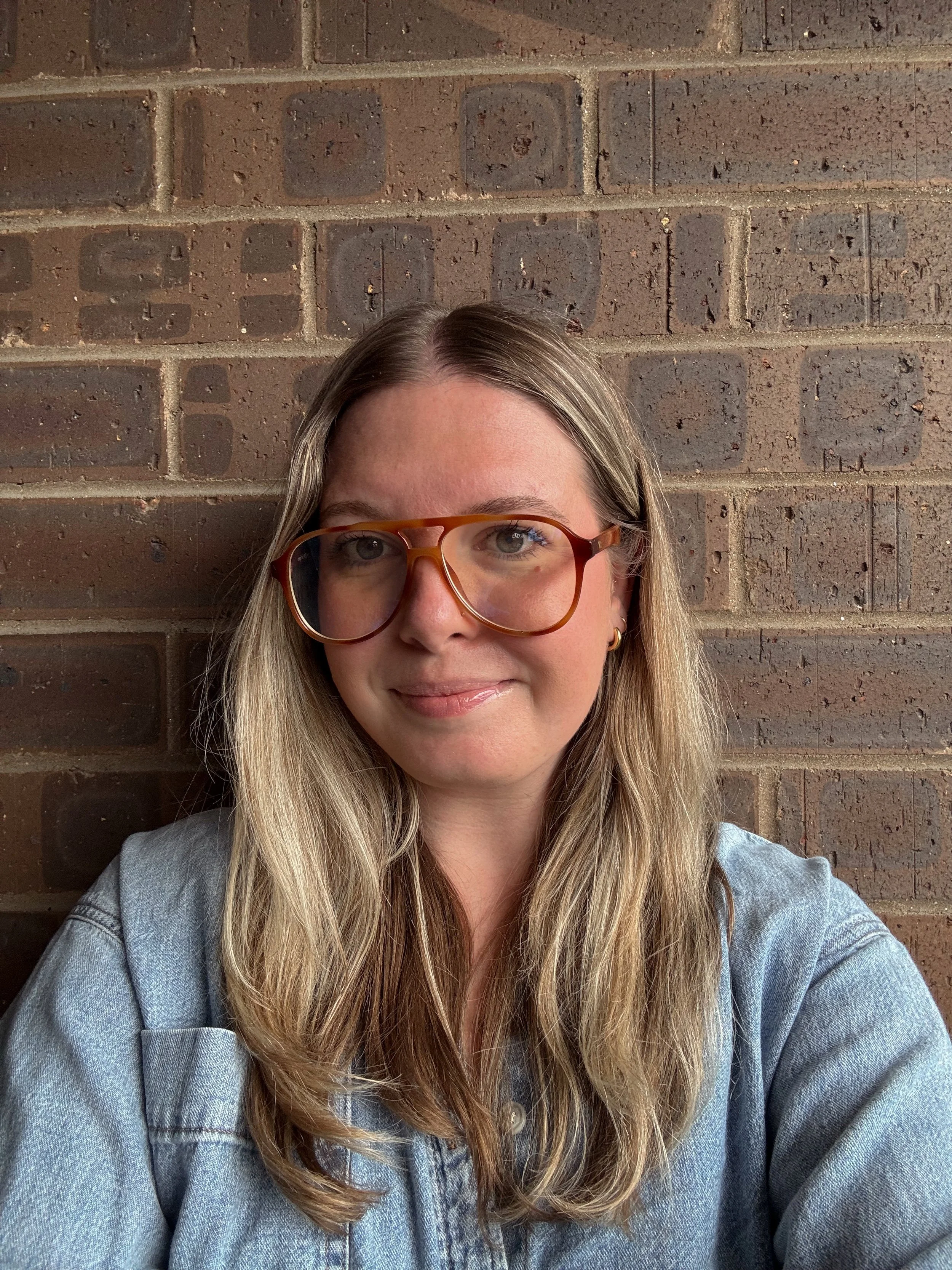 A woman with long blonde hair wearing glasses and a denim jacket, standing in front of a brick wall.