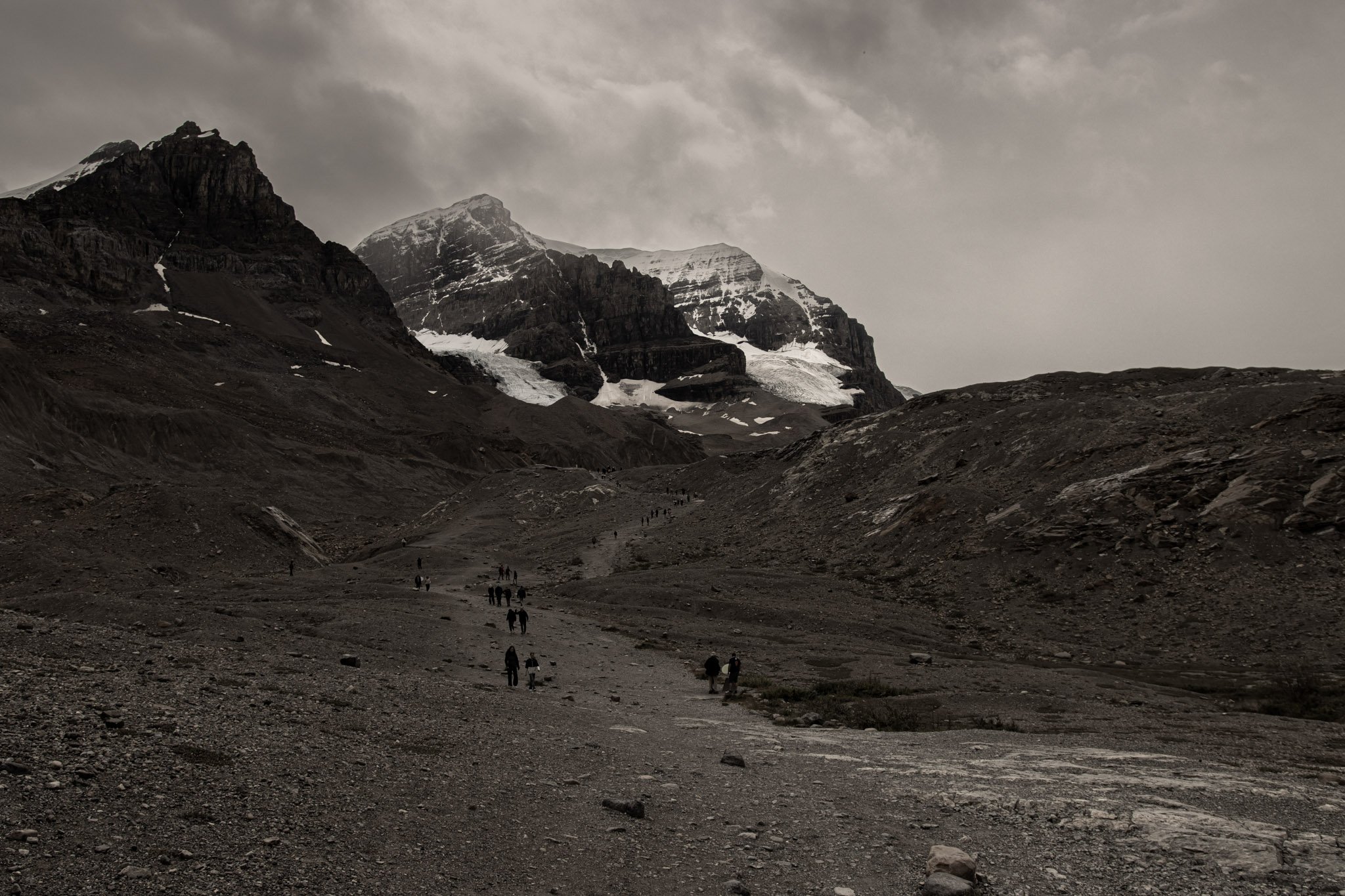 Athabasca glacier,  A mountainous landscape with dark rocky terrain, snow-capped peaks, and a cloudy sky. A group of hikers is walking along a trail in the foreground.