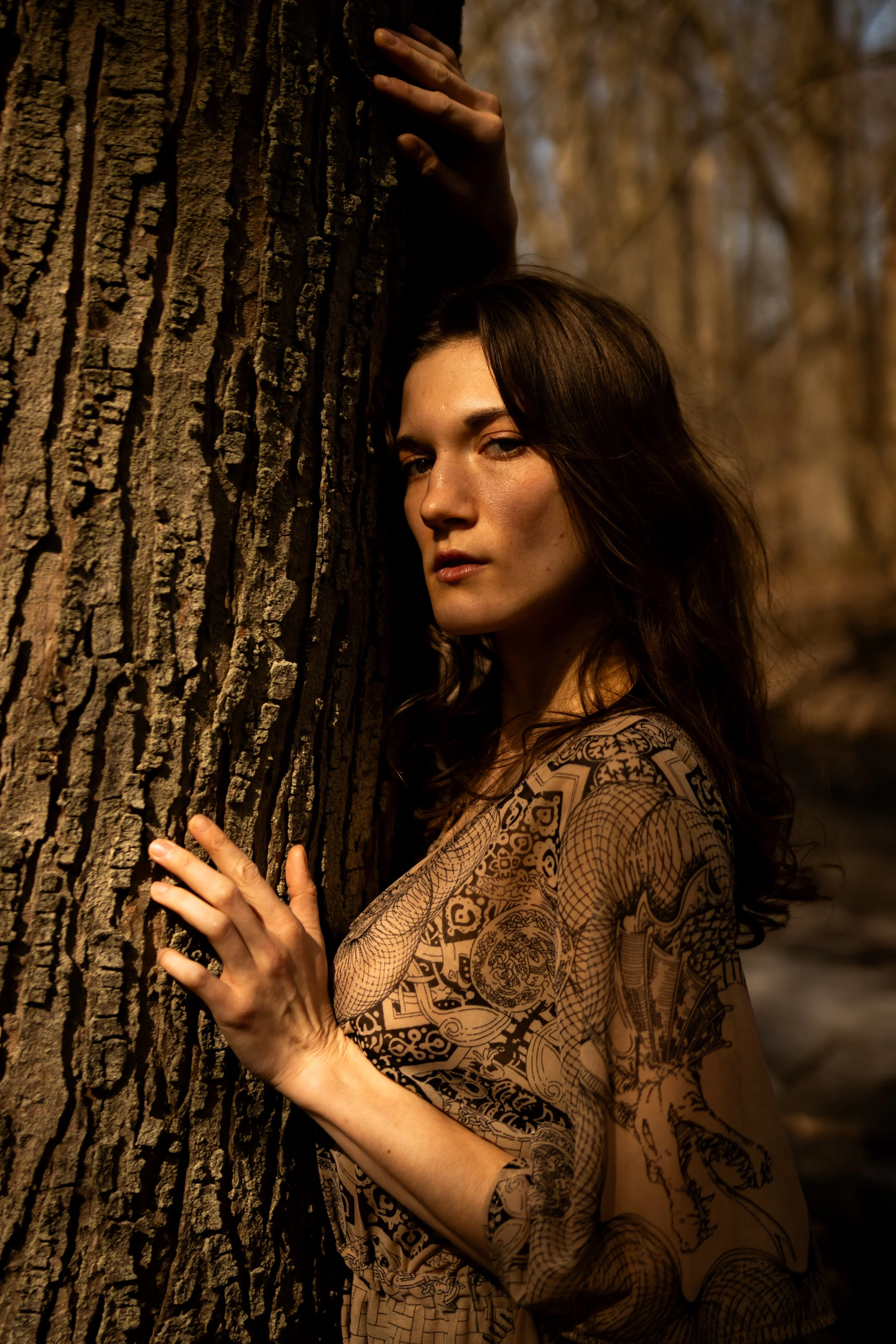 A woman with long dark hair and intricate tattoos on her arm and shoulder, standing outdoors and leaning against a tree with her hand on the bark.