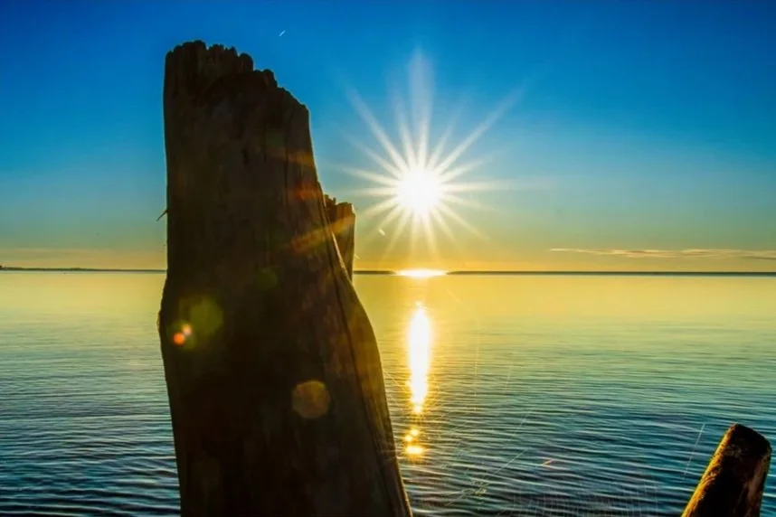 Sunset over a calm body of water with a large weathered wooden post in the foreground and the sun shining brightly in the sky.