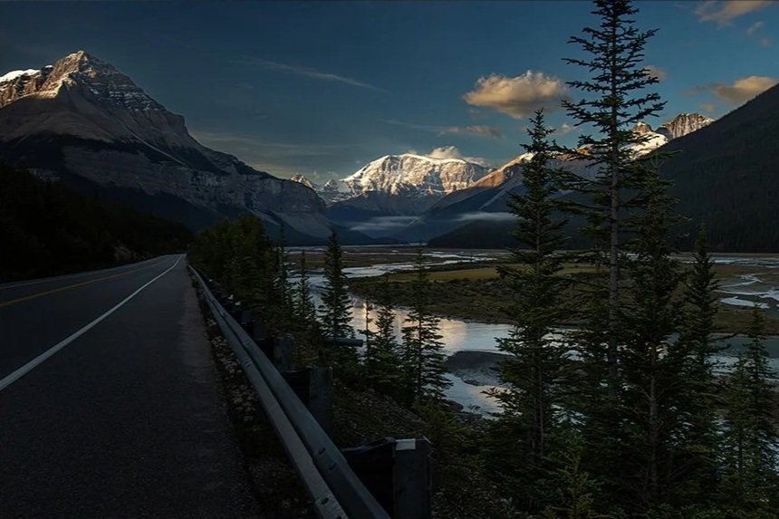 Scenic mountain landscape with a road on the left, tall pine trees in the foreground, a river, and snow-capped mountains under a partly cloudy sky.
