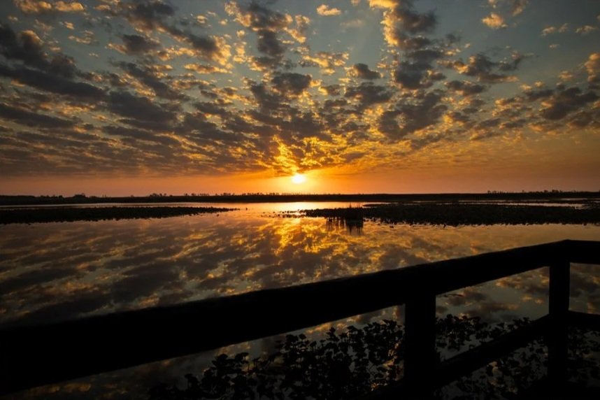 Sunset over a body of water with a cloudy sky reflected on the surface, viewed from a wooden railing at a lakeside or riverside location.