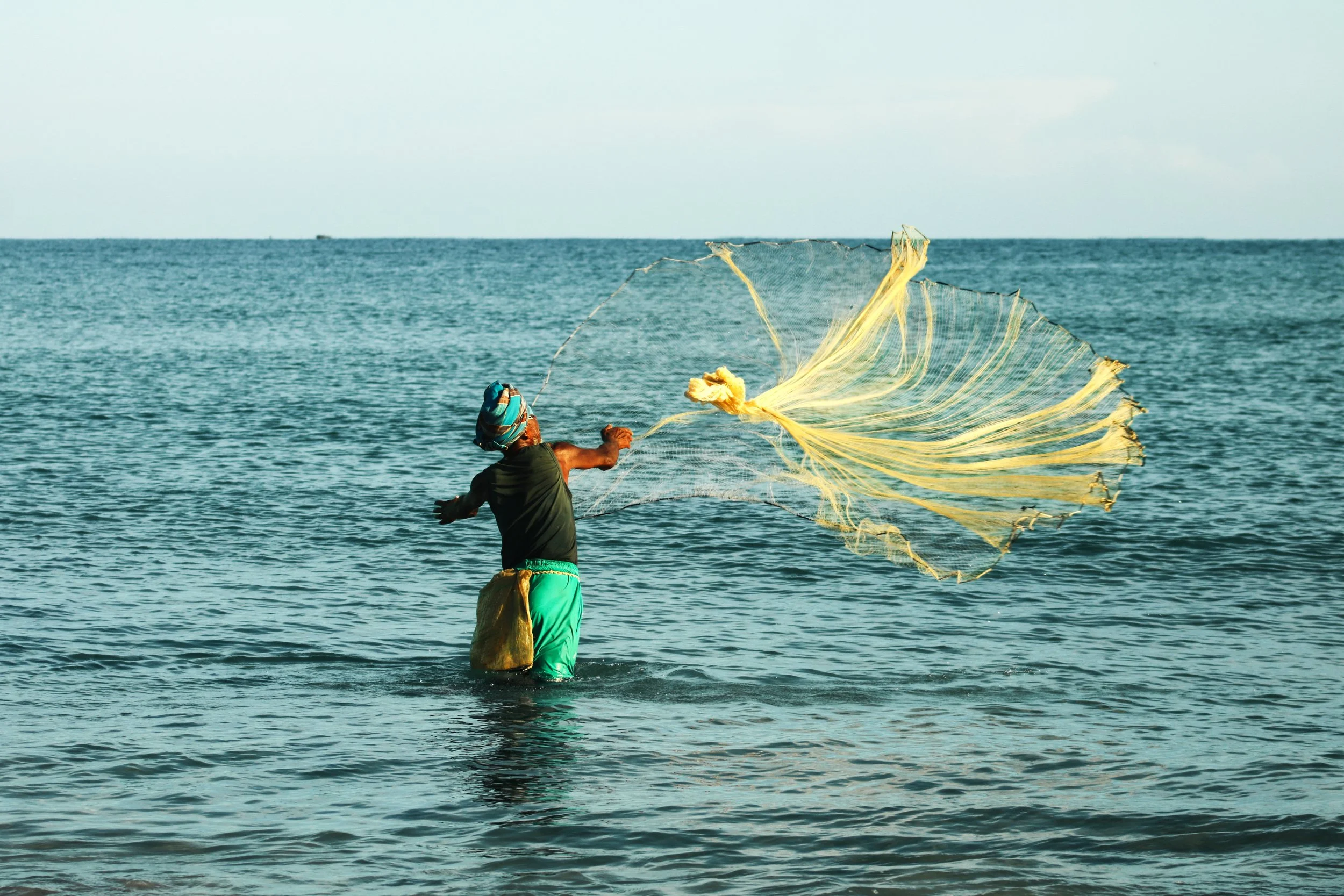 Fisherman wearing a colorful head wrap and green pants casting a fishing net into the ocean.