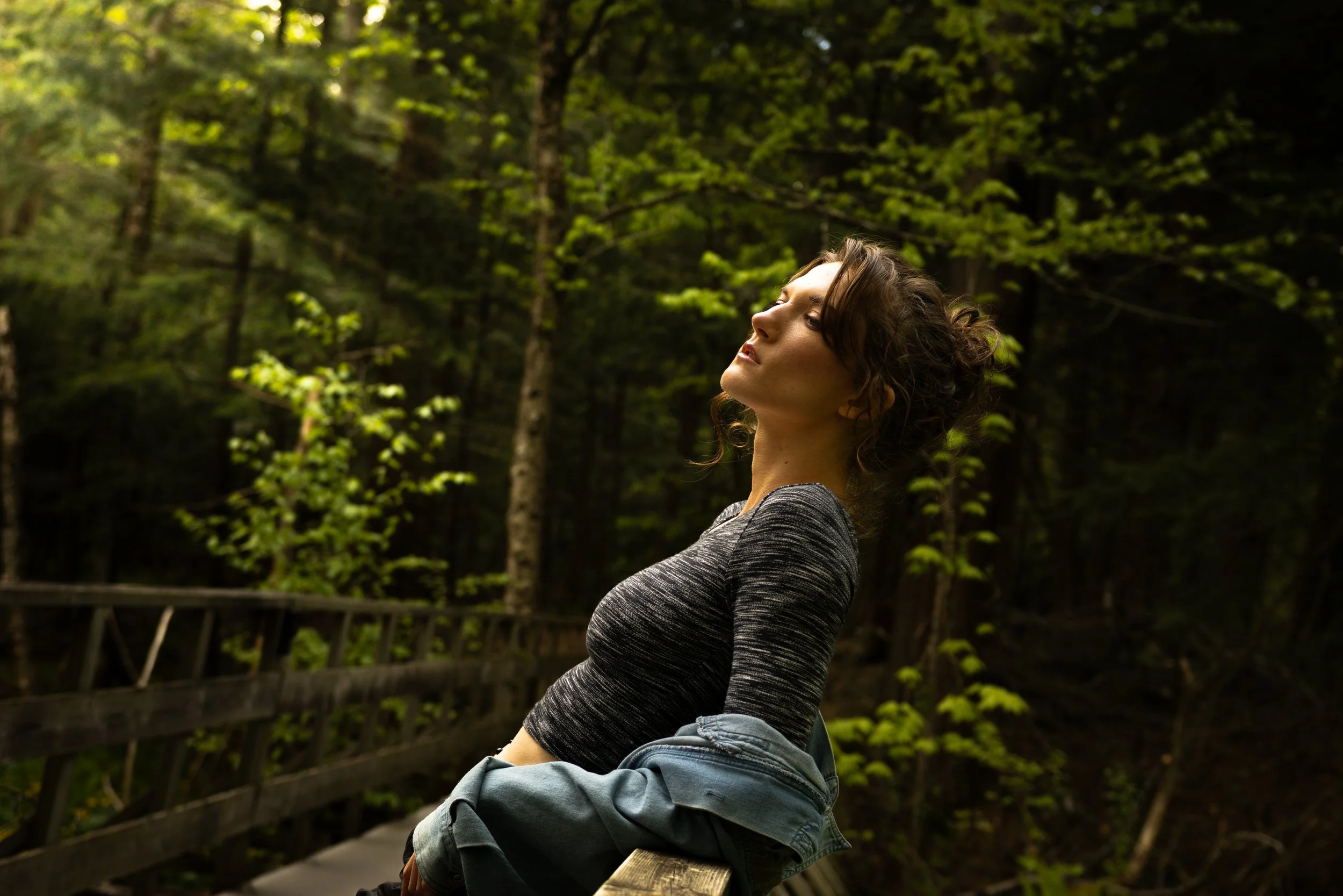 A woman with curly hair sits in a green forest with her eyes closed and head tilted back. Moody Toronto portrait photography by Dark Art Photos.