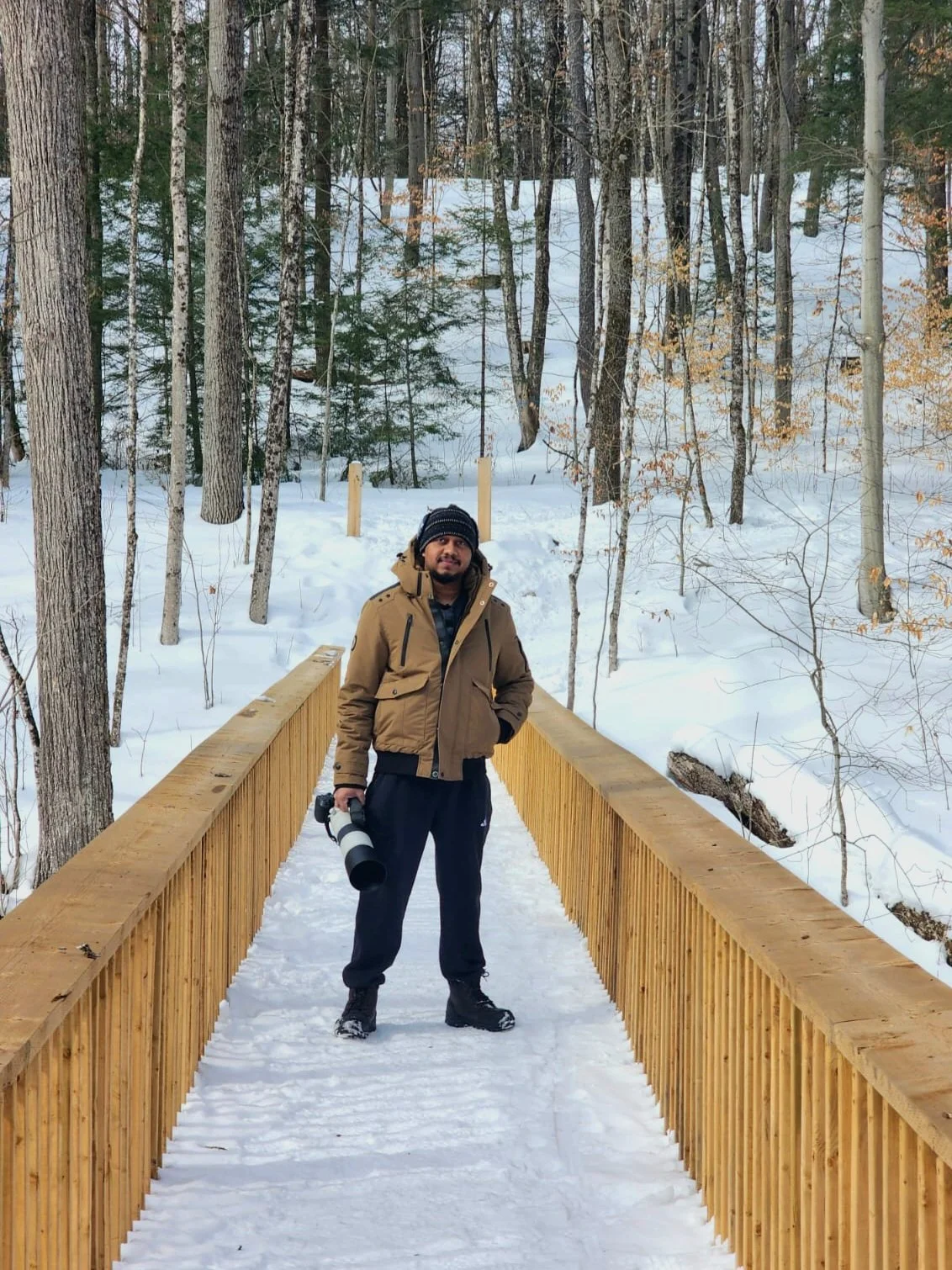 Owner Dark Art, stands on a wooden bridge in a snow-covered forest, holding a camera with a large lens, wearing a brown jacket, black pants, and a black knit cap.