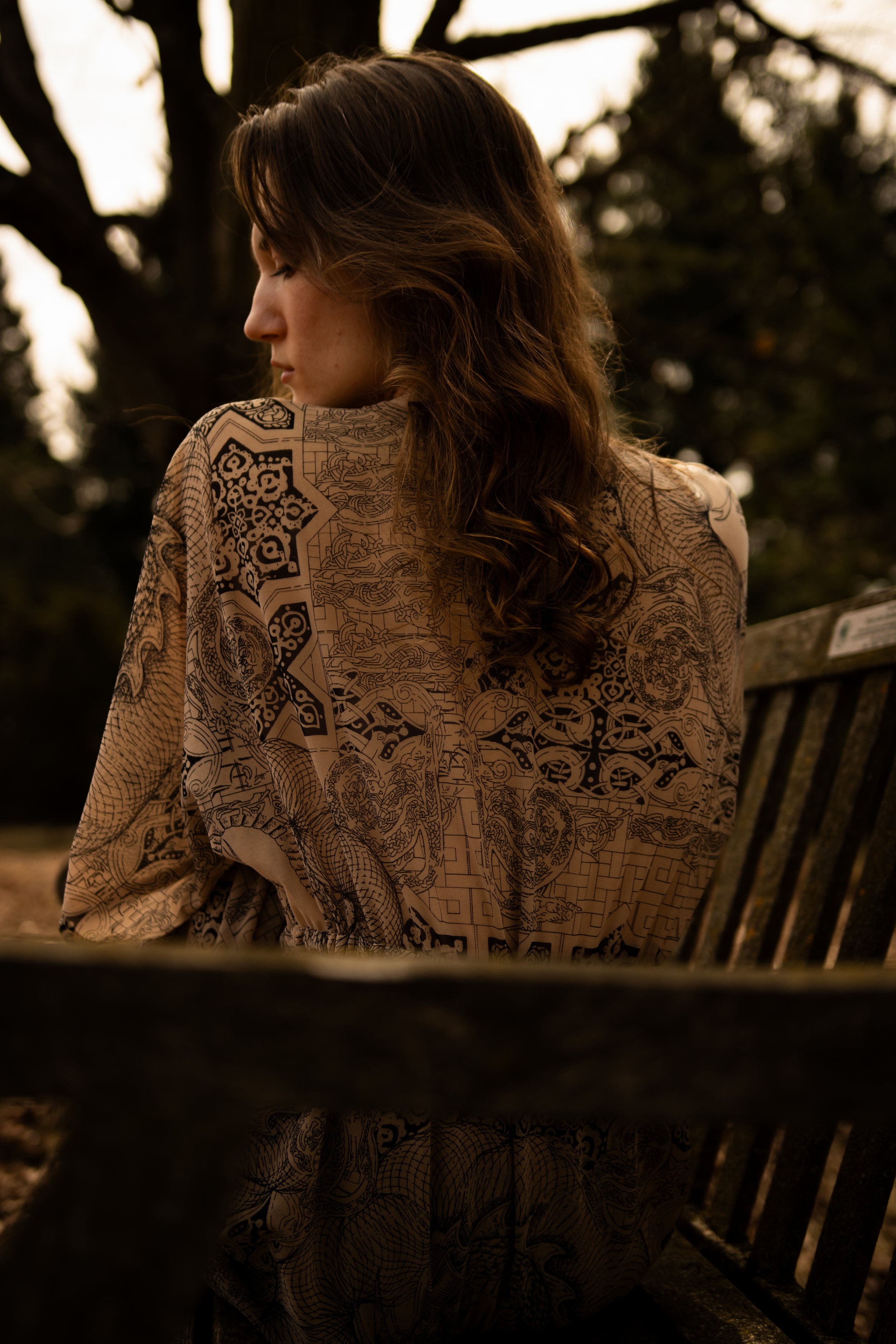 A woman with wavy brown hair sits on a wooden bench at sunset in this high-contrast outdoor portrait. Shot by Toronto photographer Dark Art Photos.