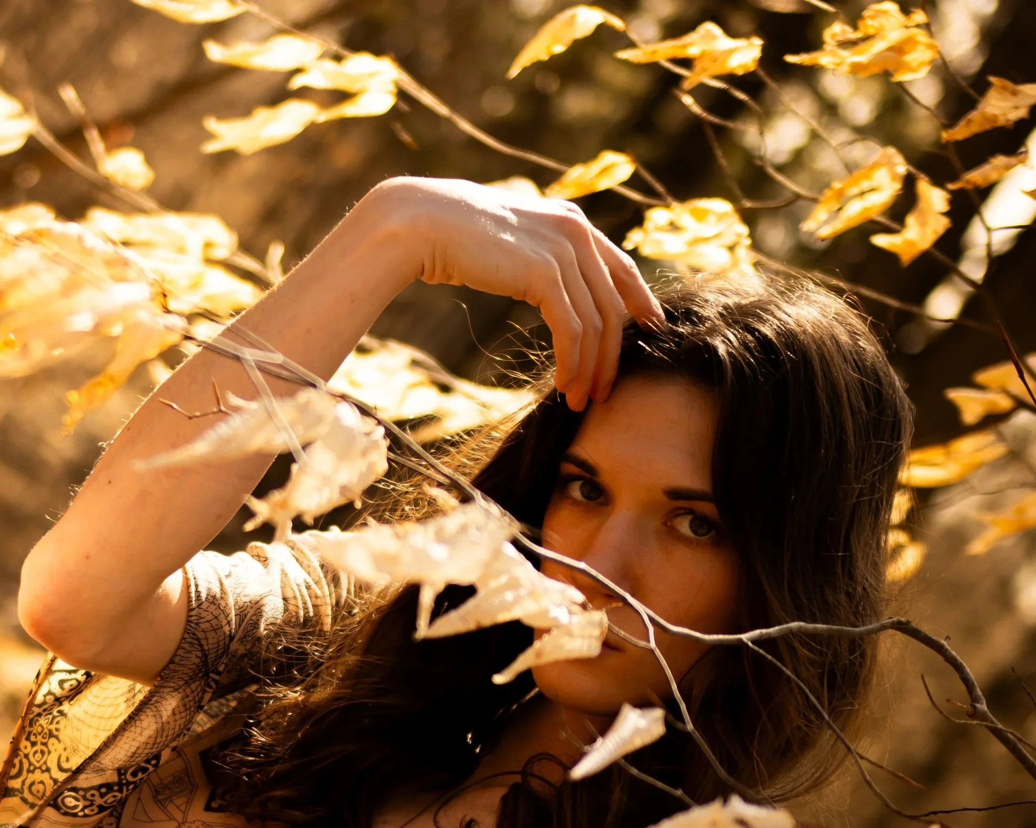 A woman with dark hair and tan skin is outdoors among dry, yellow leaves. She has one hand resting on her head, and she is partially visible with her face peeking out from behind the leaves and branches.