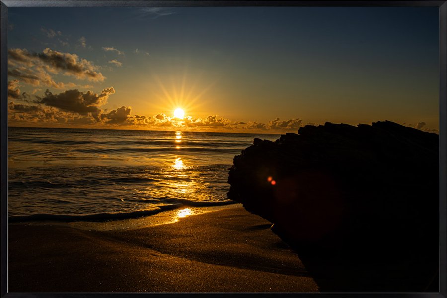 Sunset over the ocean with waves hitting the sandy beach and rocks in the foreground.
