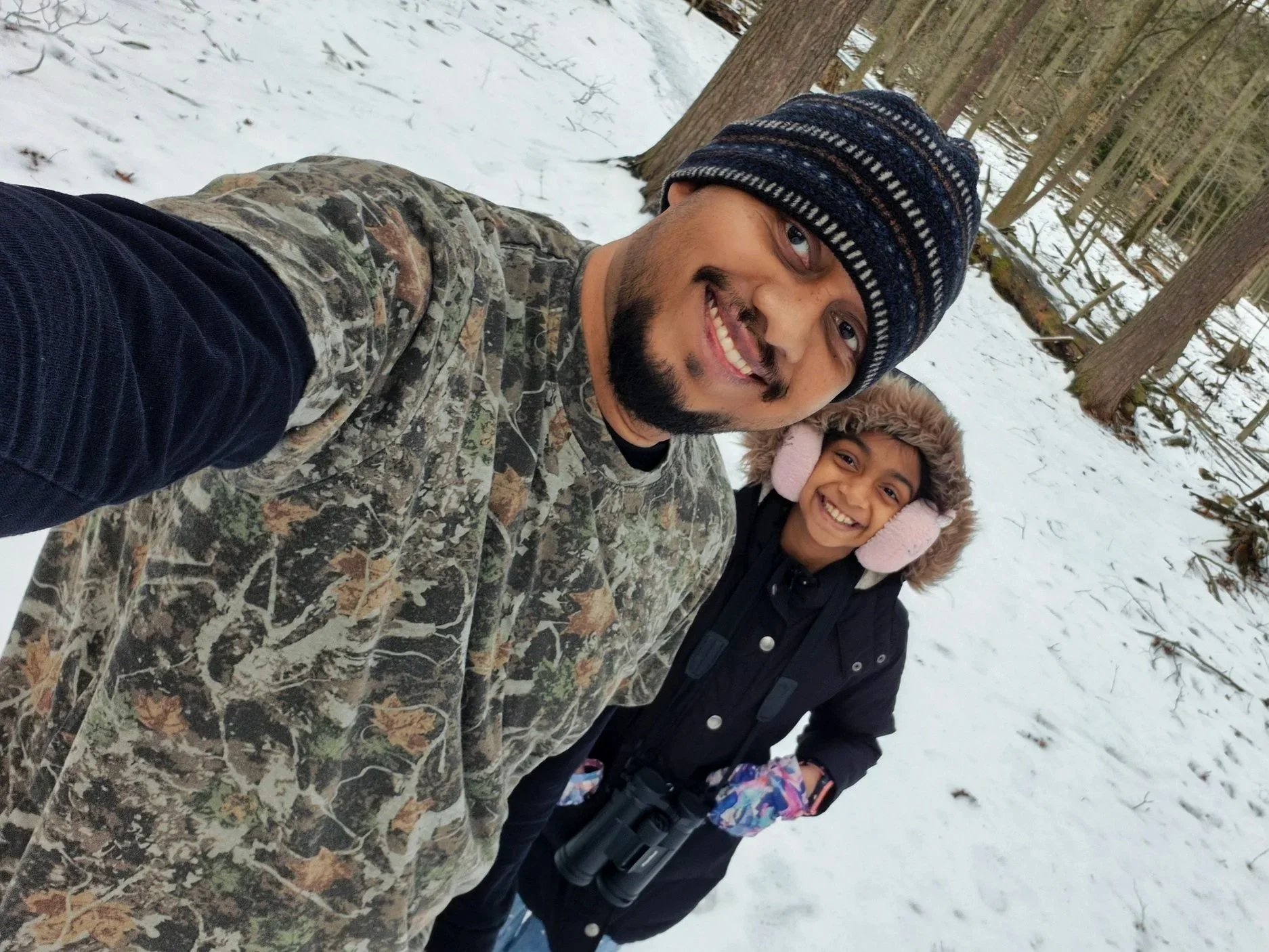 Two smiling individuals taking a selfie in a snowy forest (Owner Dark Art and his Daughter), one wearing a camouflage jacket and a blue knit hat, the other wearing a dark coat and pink earmuffs.