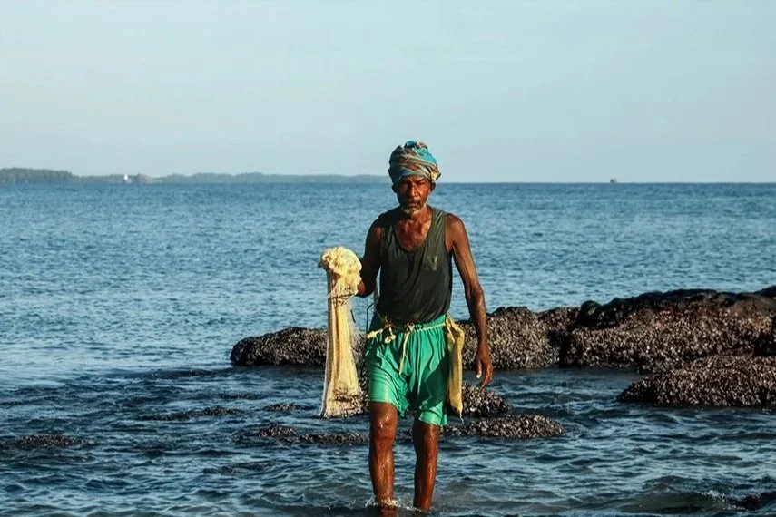 A man stands in the shallow ocean water near rocks, wearing a turban, a sleeveless shirt, and green shorts, holding a net with a shell on it.