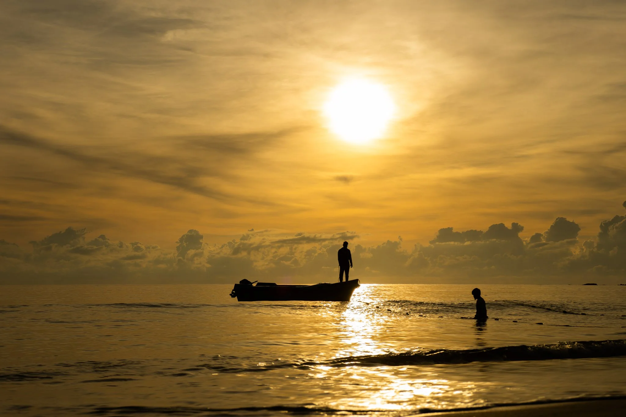 A person standing on a small boat on the water during a golden sunset, with clouds in the sky and another person in the water nearby.