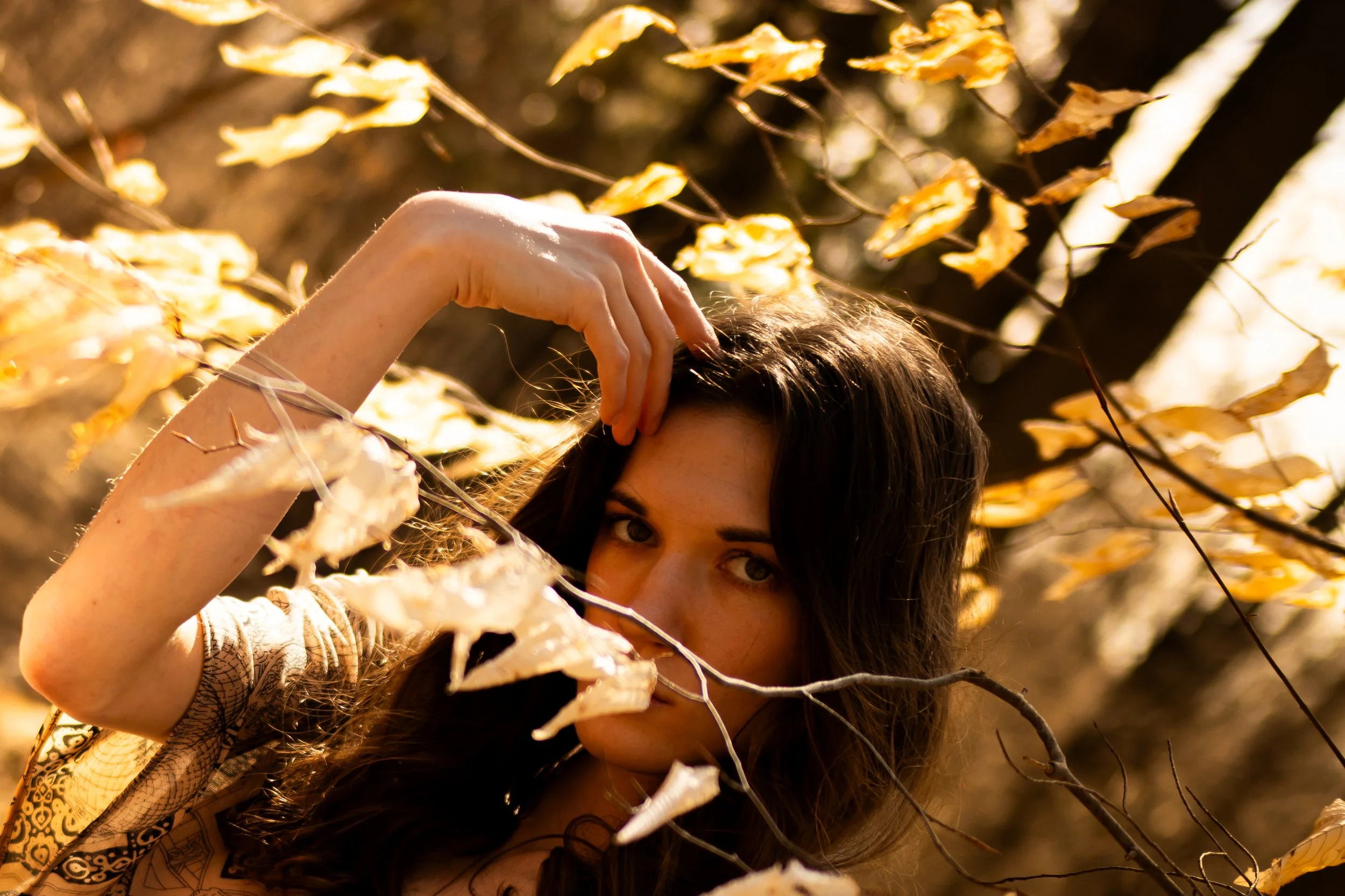 A woman with long dark hair peeking through autumn leaves. High-contrast, moody portrait photography in Toronto by Dark Art Photos.