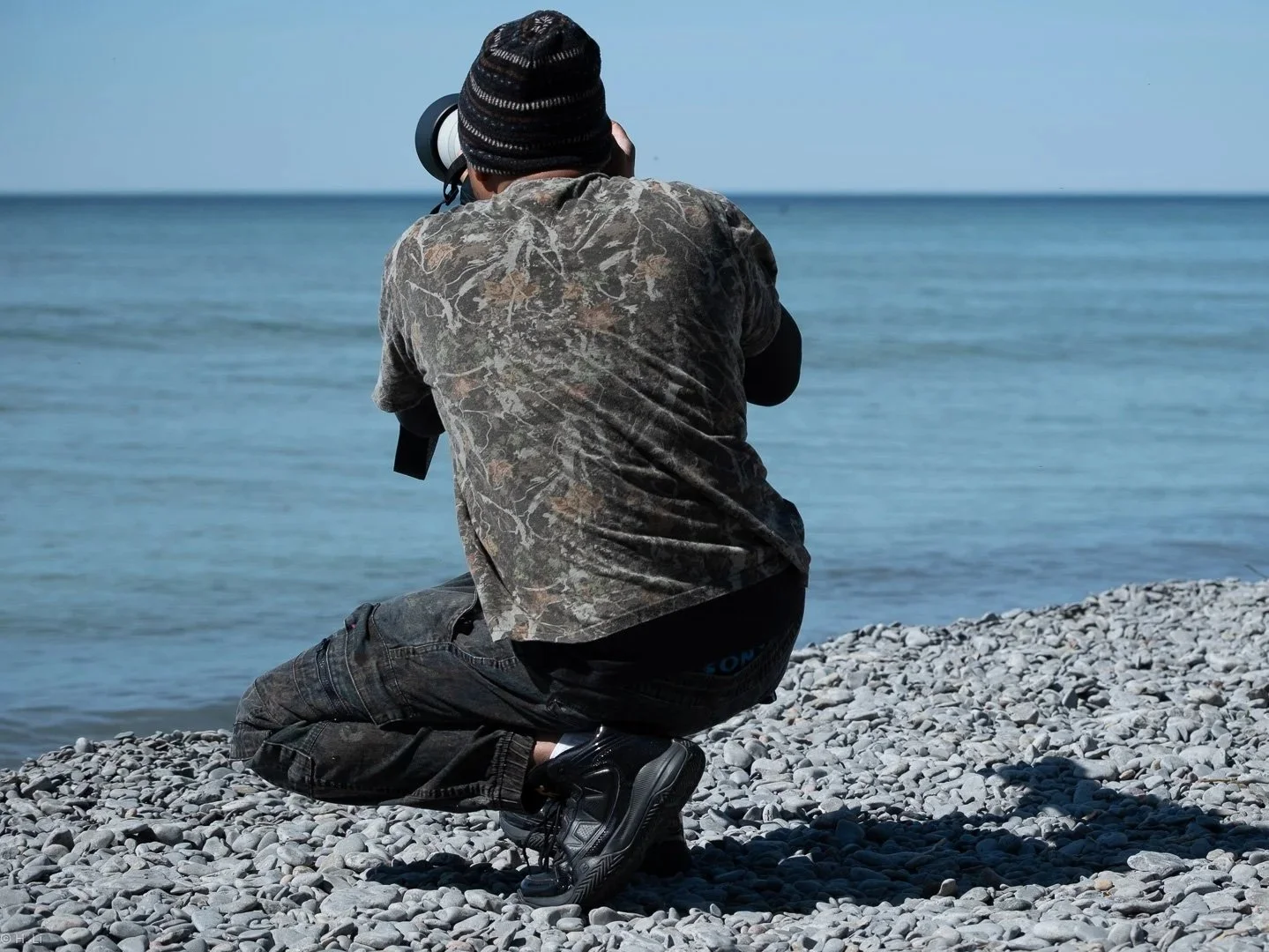 Owner Dark Art, kneeling on a rocky beach, aiming a camera at the ocean, with the horizon and clear sky in the background.