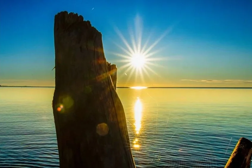 Sunset over calm lake with a wooden post in the foreground and the sun reflecting on the water.