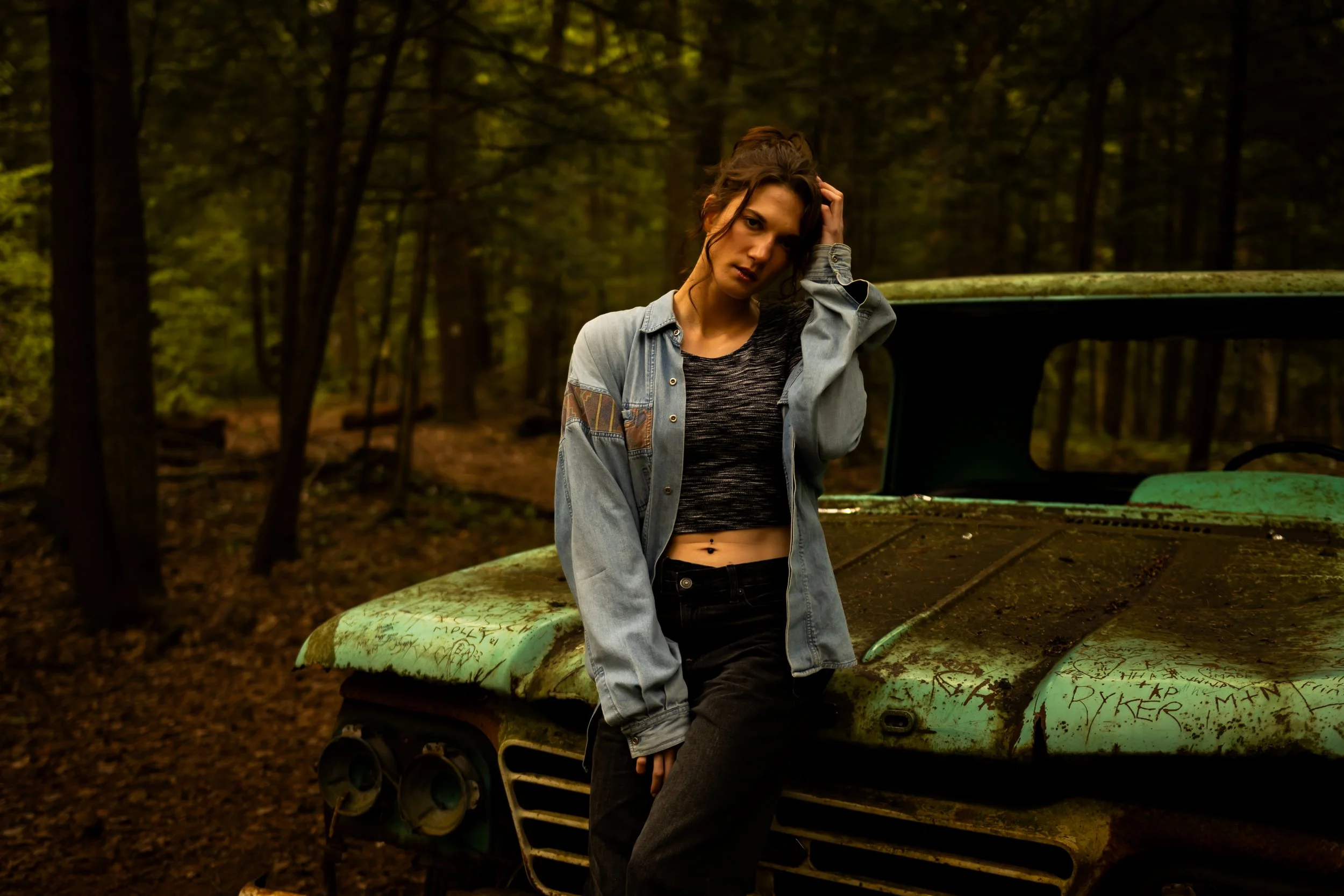 Cinematic portrait photography by Dark Art Photos in Toronto. A young dark-haired woman poses near a vintage, graffiti-covered green car in a moody wooded area.