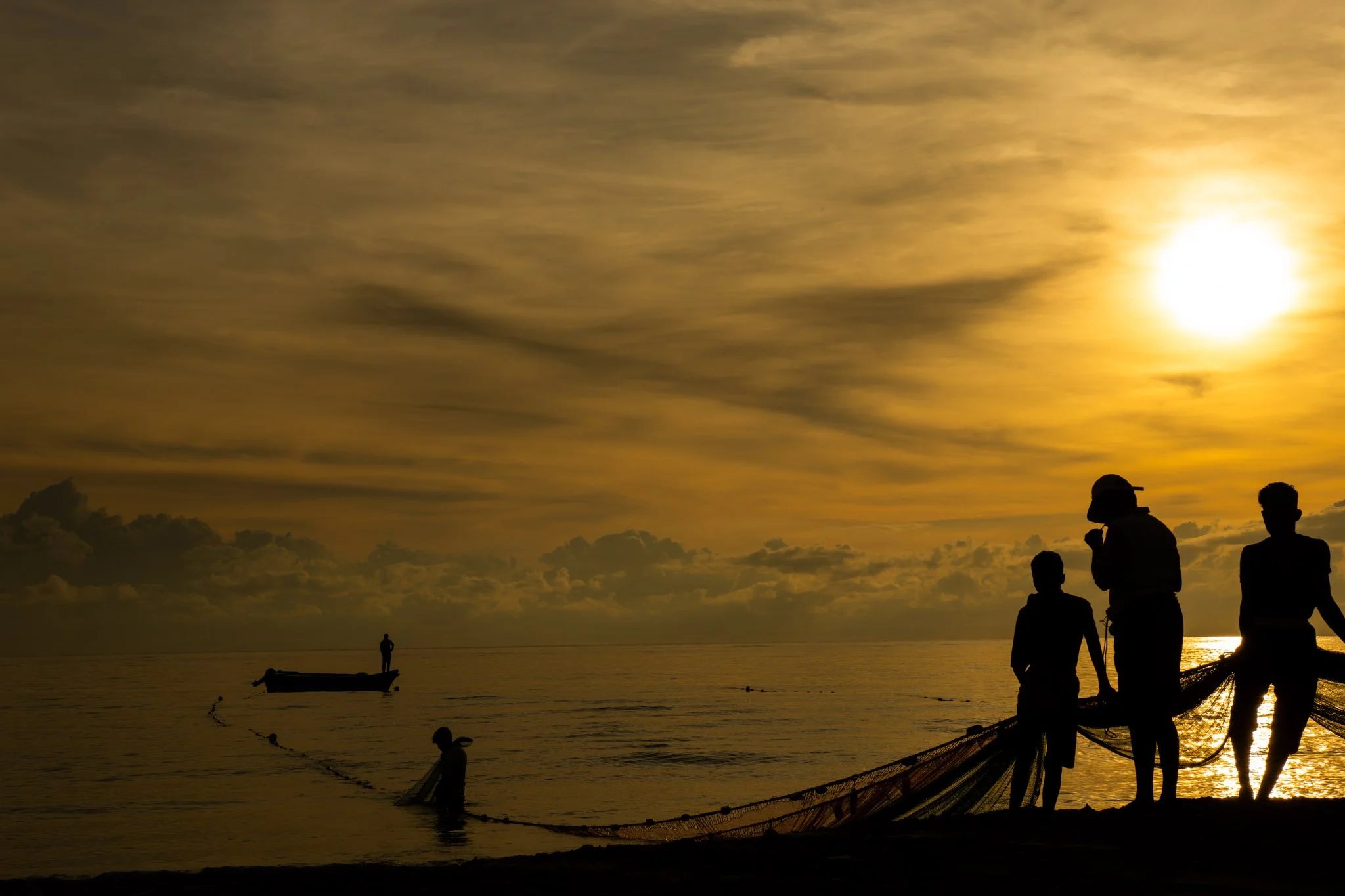 Silhouettes of Fisherman at the beach during sunset, some fishing and others standing with fishing nets, with a boat in the water and a partly cloudy sky illuminated by the setting sun.