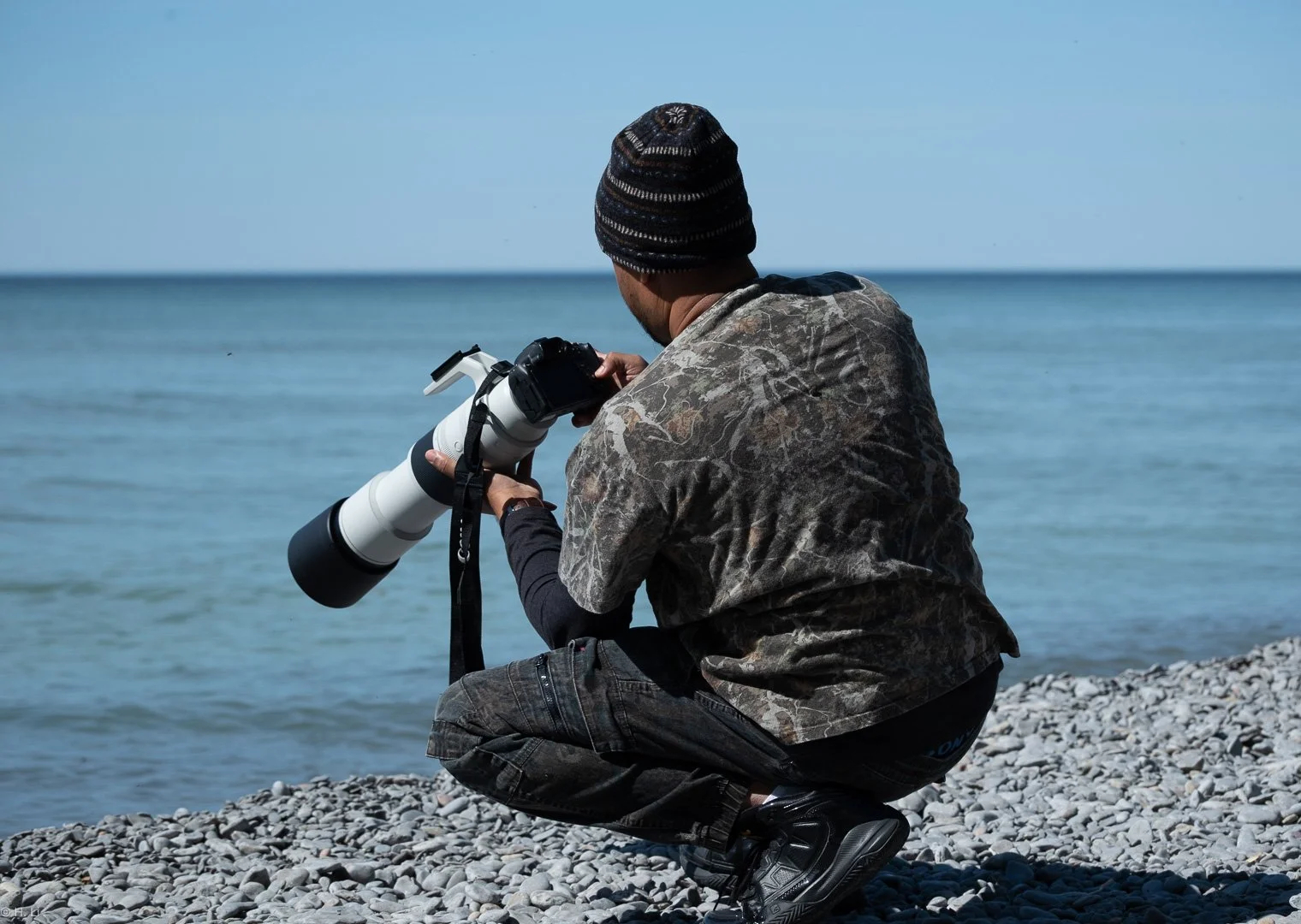 Owner Dark Art, crouching on a rocky beach by the water, looking through a camera with a long telephoto lens.