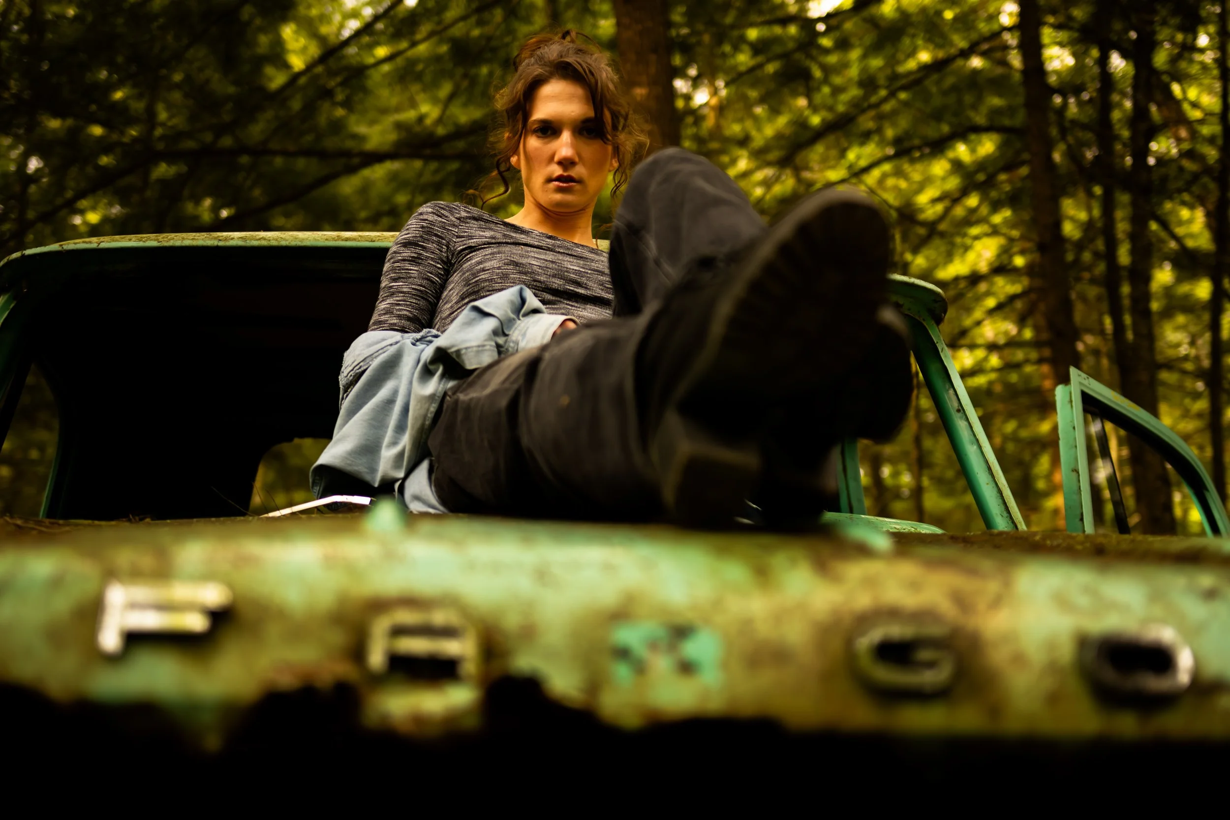 Dark-toned fine art portrait of a woman looking downward while lying on an old green tractor in a forest. Cinematic Toronto photoshoot by Dark Art Photos.