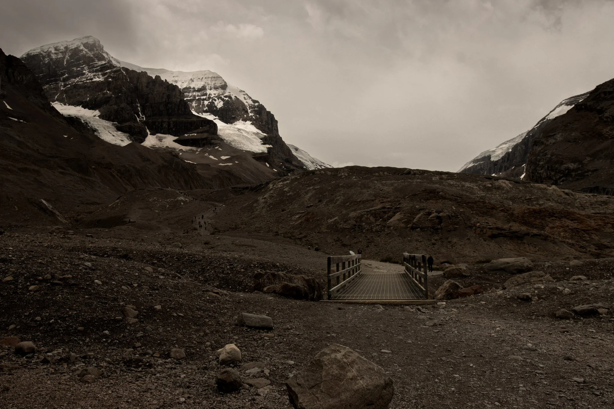 Athabasca glacier, A rugged mountain landscape with snow-capped peaks and rocky terrain under a cloudy sky, featuring a small wooden bridge and a few hikers.