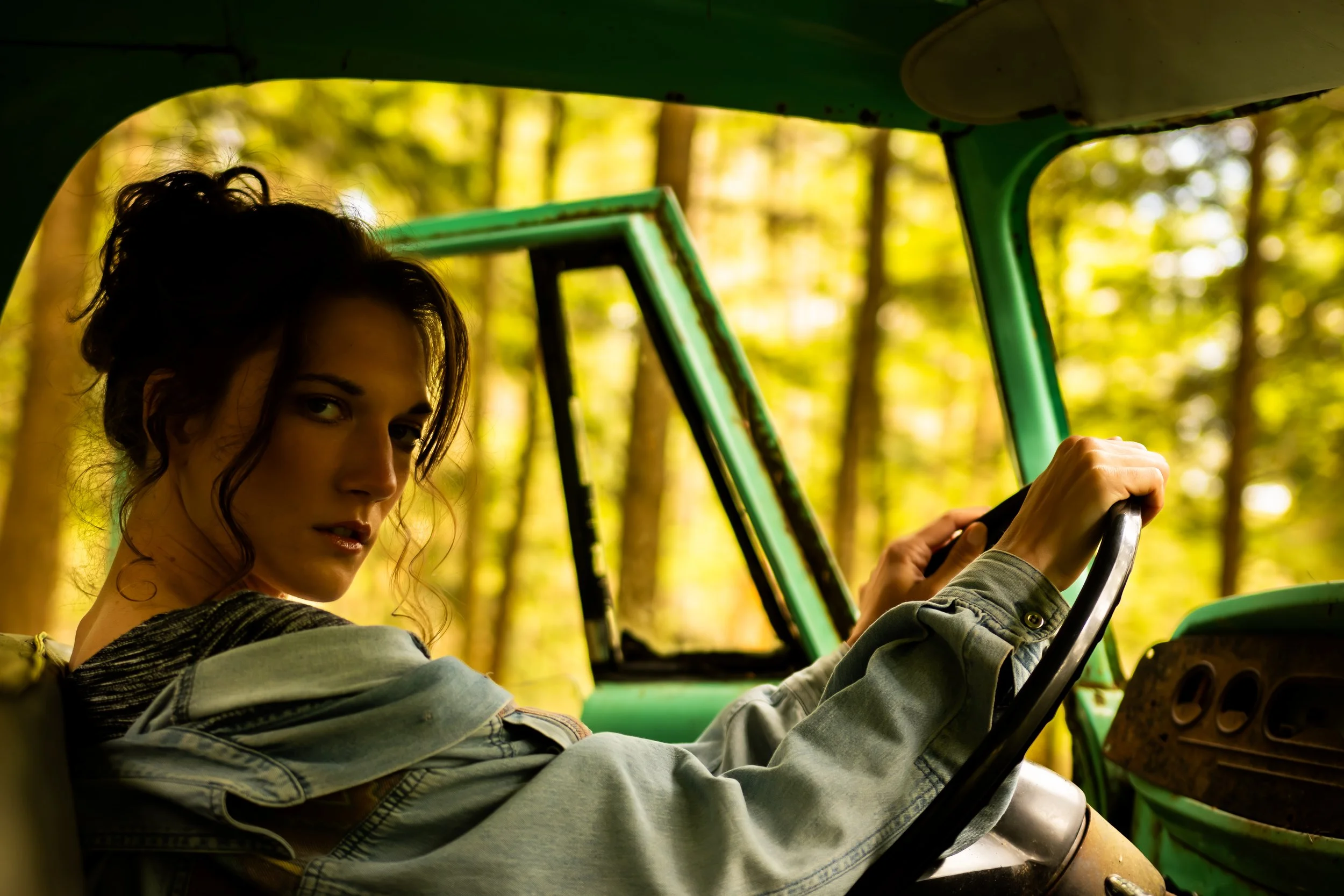 A woman with curly hair driving a vintage green vehicle through a forest with sunlight filtering through trees.