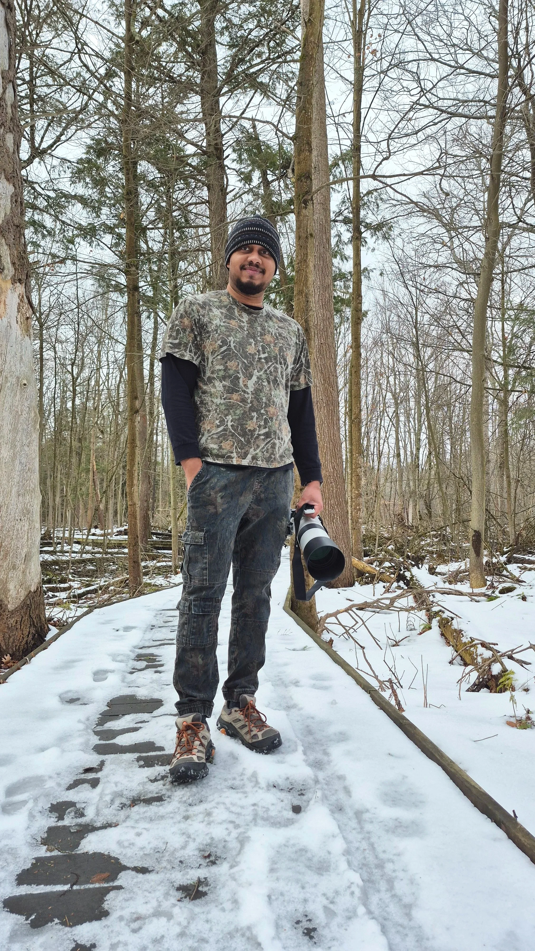 Owner Dark Art, in winter clothing, camouflage shirt, beanie, and hiking boots stands in a snowy forest holding a camera with a long lens.