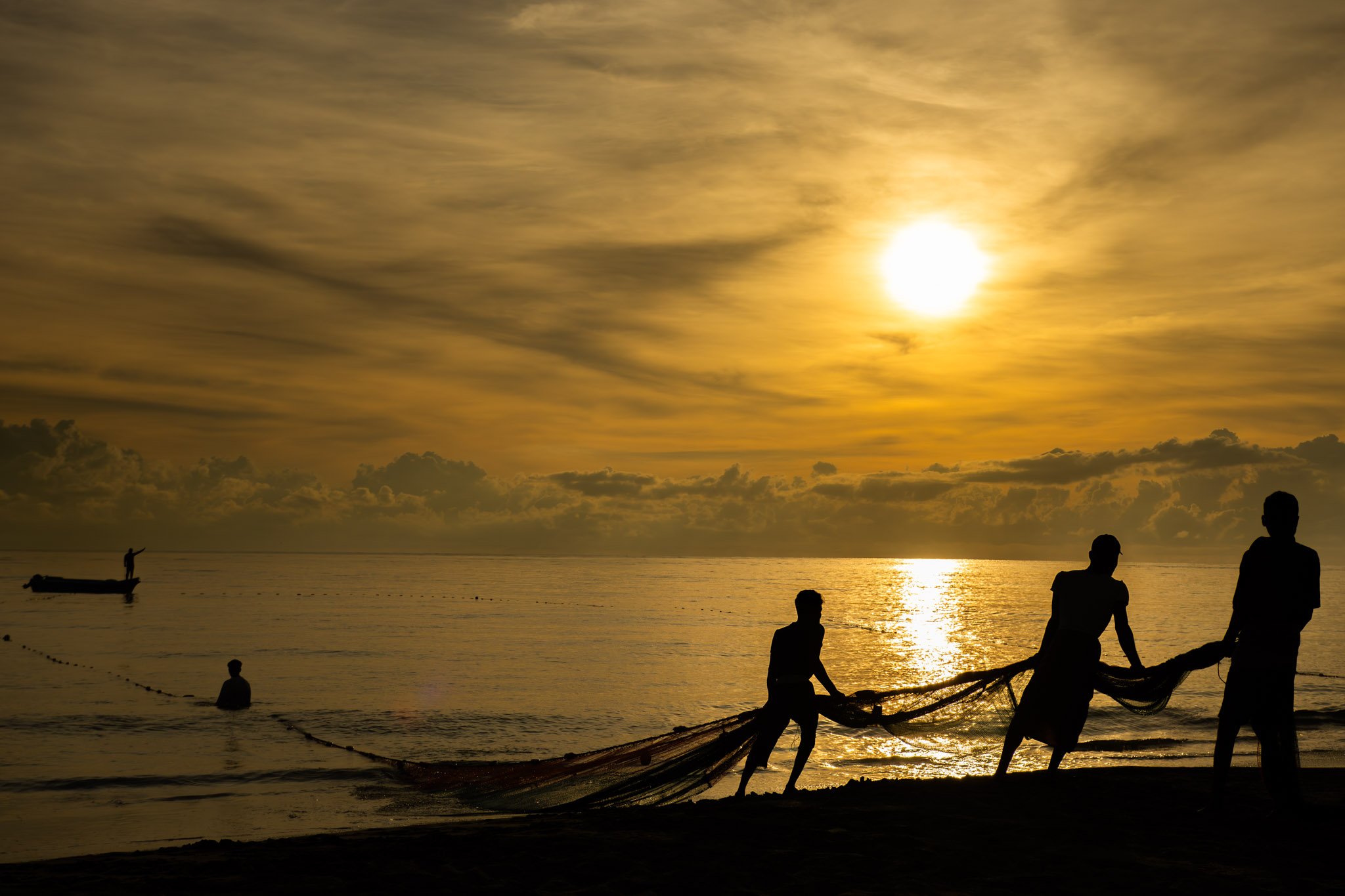 The image depicts a group of silhouetted Fisherman at the beach during sunset, with a large, bright sun, partly cloudy sky, calm water, and a small boat with a person standing in it.