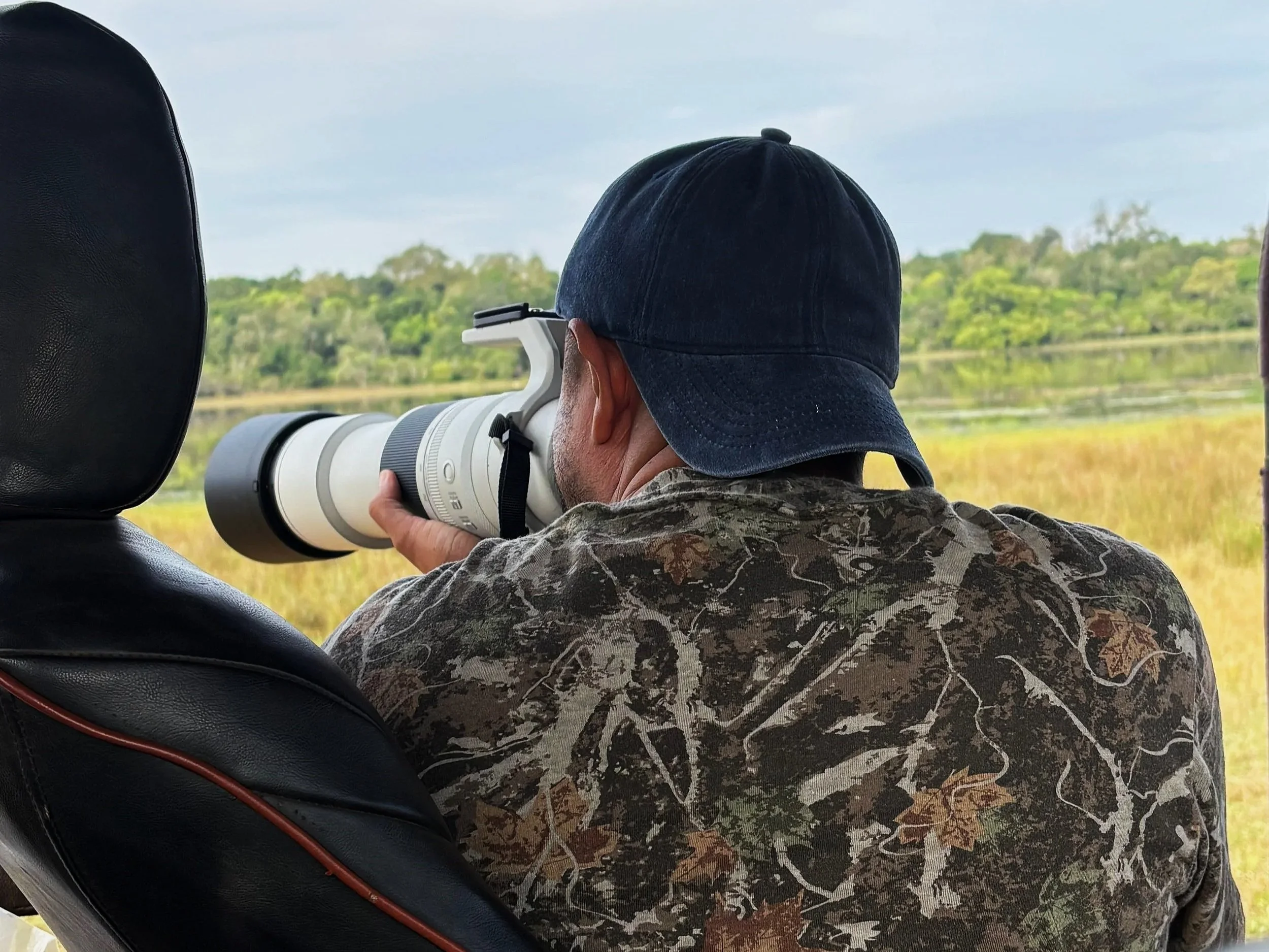 Owner Dark Art, wearing a dark cap and camouflage clothing, sitting in a vehicle, looking through a telescope or camera, with a landscape of trees and water in the background.