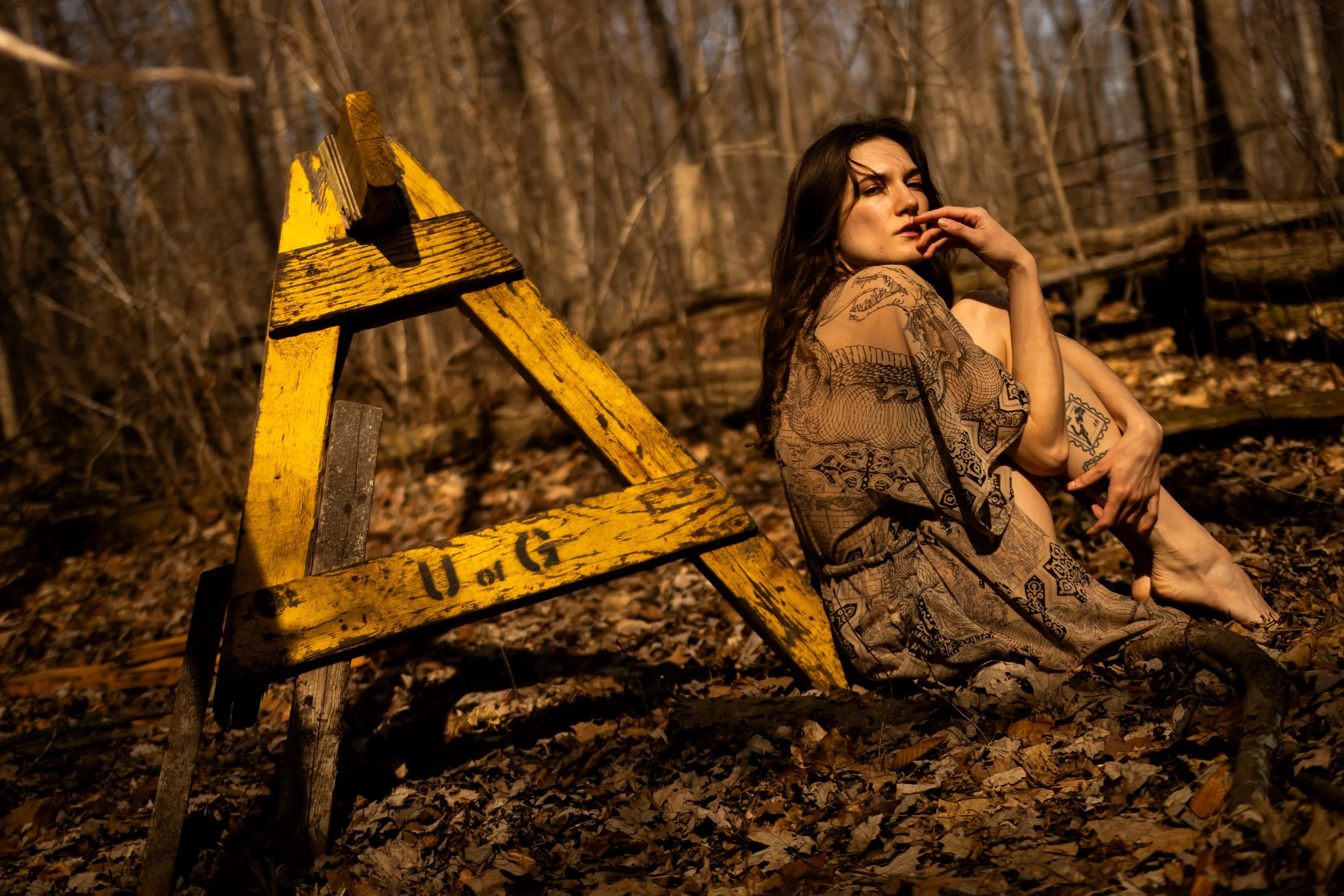 A woman with long dark hair and tattoos sitting outdoors in a wooded area, touching her face with one hand, beside a yellow wooden barricade with black markings, during a sunny day.