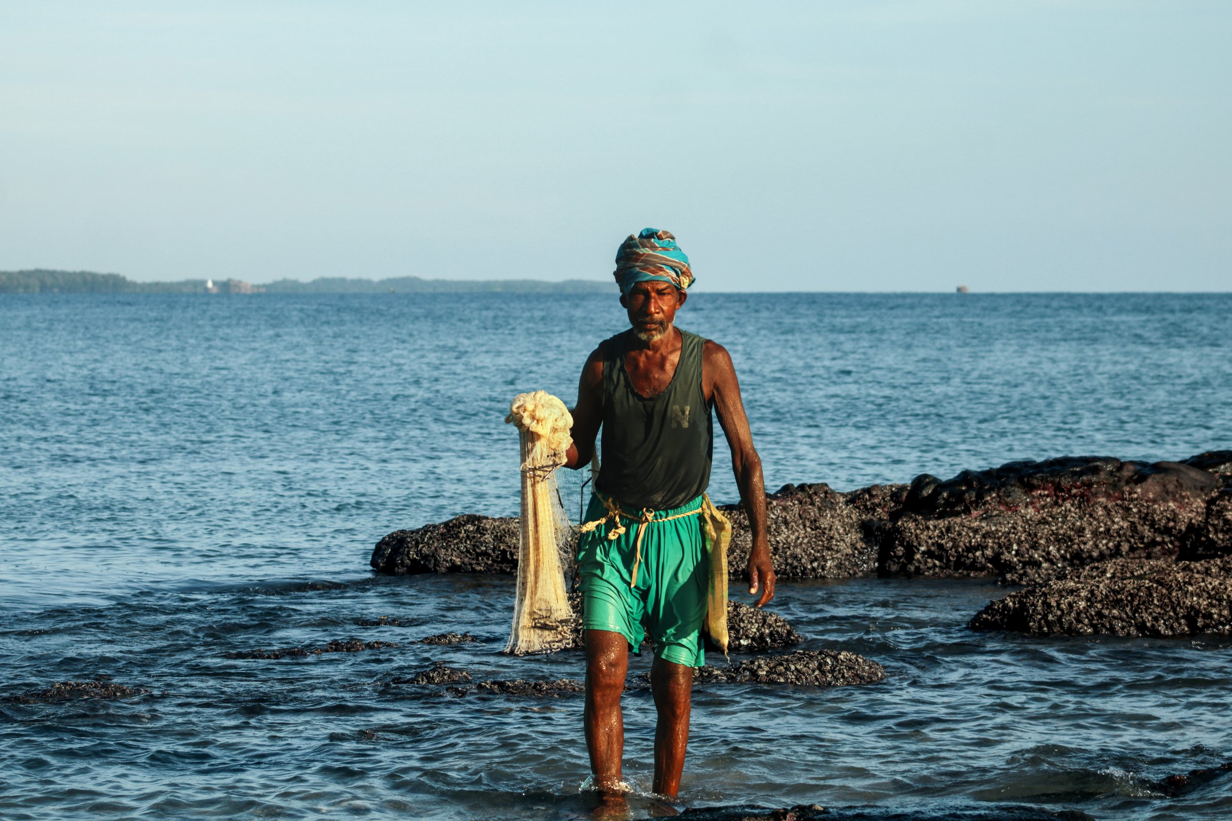 An elderly man with a colorful head wrap walking out of the water carrying a fishing net, with rocks and the ocean in the background during daytime.