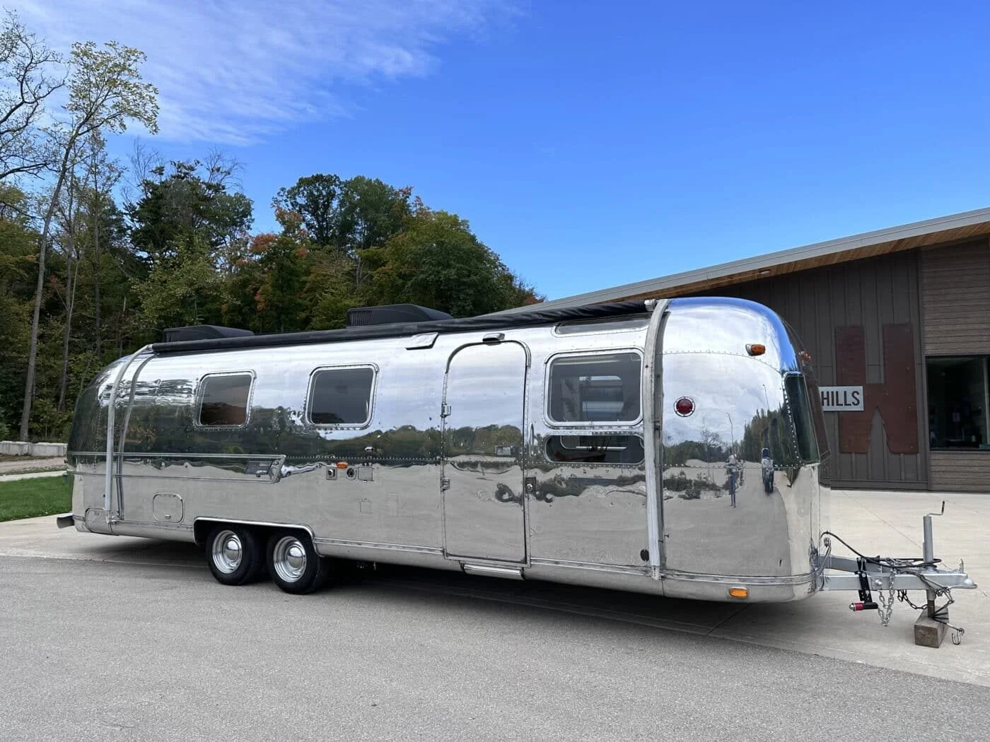 A shiny, silver Airstream travel trailer parked on a paved lot near a wooden building with a sign that says 'HILLS.' Surrounding trees with autumn foliage and a clear blue sky are in the background.