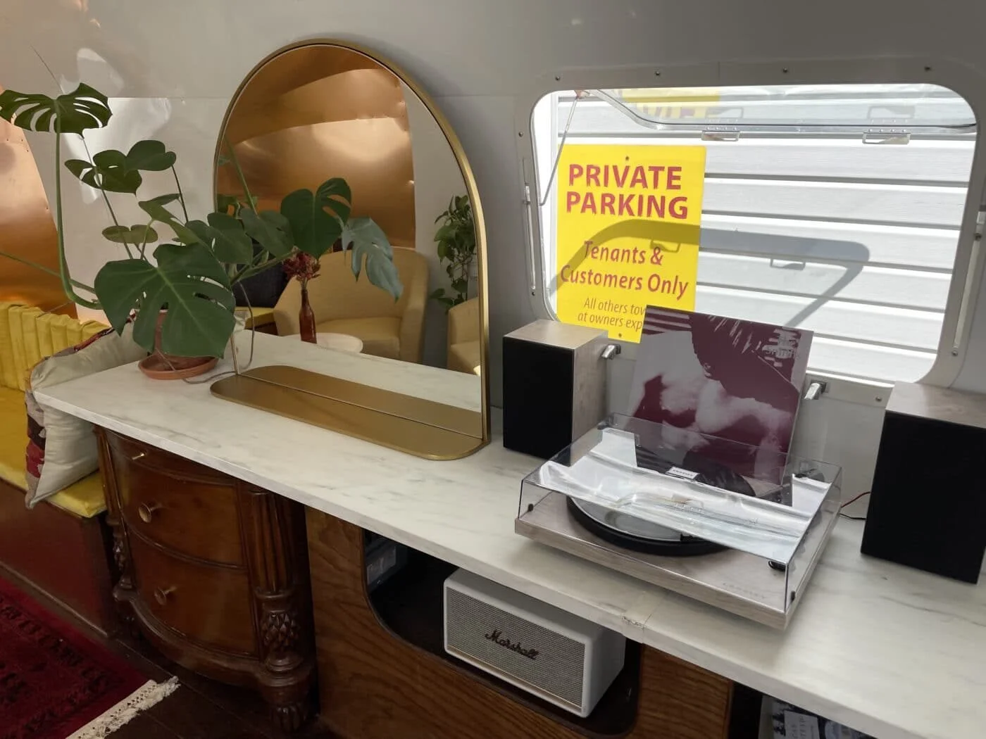 Interior of a vintage trailer with a marble-topped dresser, green plants, a turntable, and a sign indicating private parking for tenants and customers.