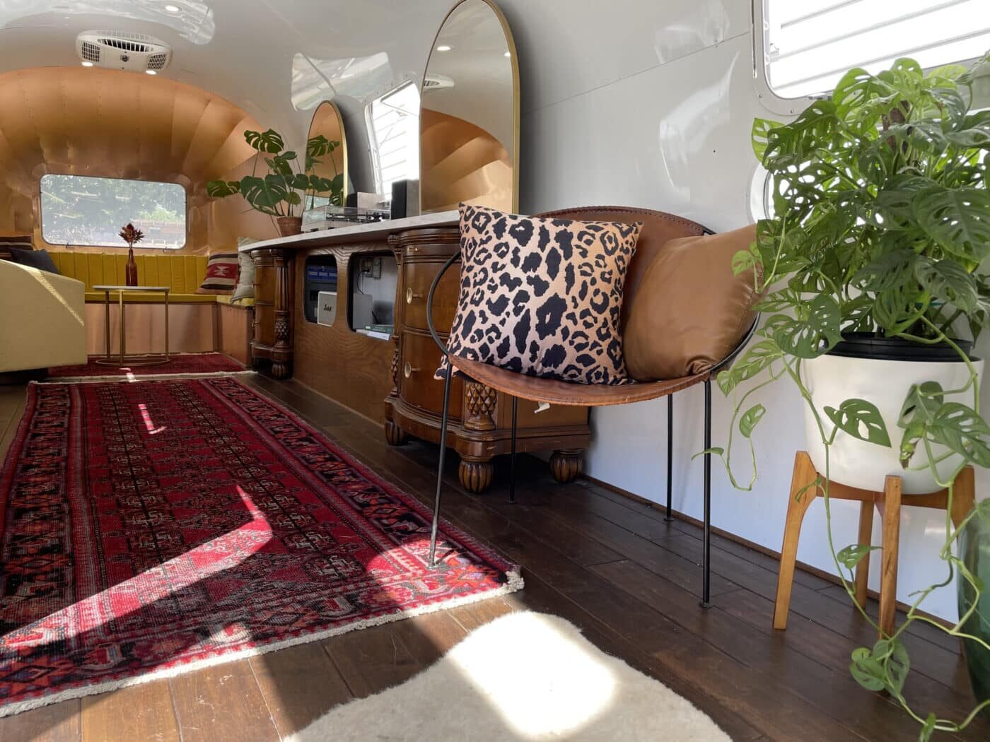 Interior of a cozy camper with a red patterned rug, a wooden cabinet, a large mirror, potted plants, a beige armchair with a leopard print pillow, and a window letting in natural light.