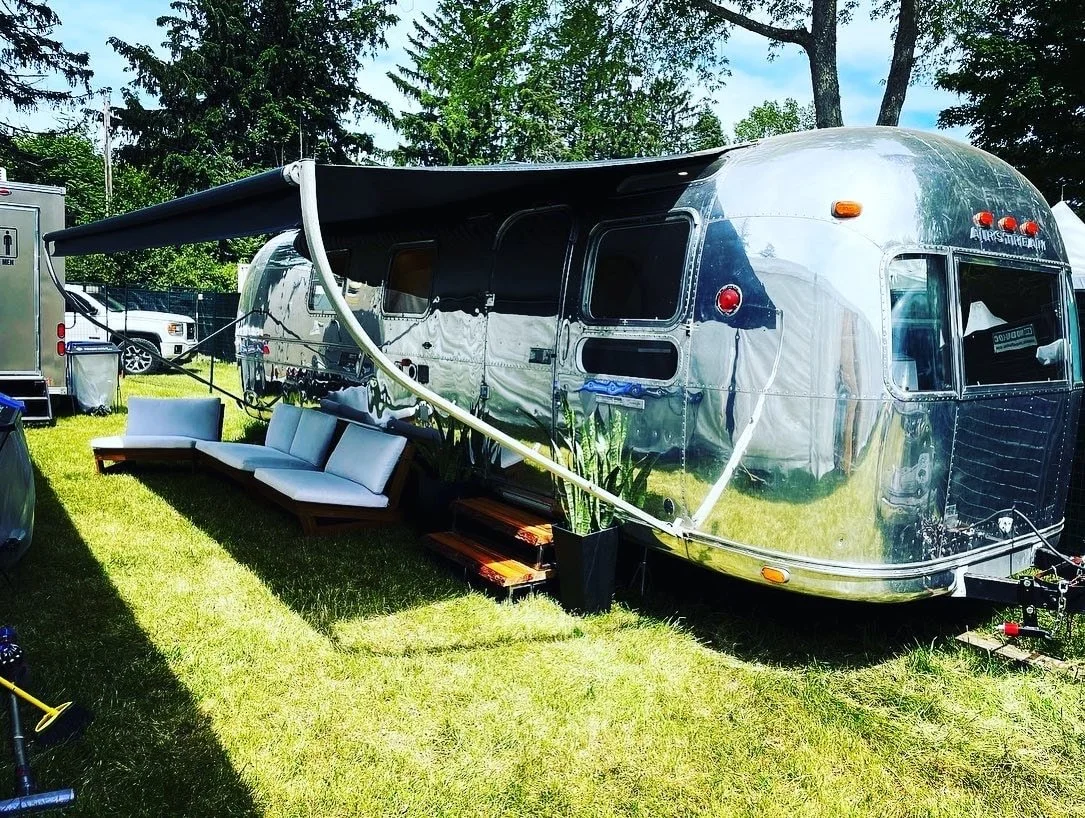 A shiny, metallic travel trailer with a black awning extended, outdoor seating with white cushions, and potted plants on a grassy area with trees in the background.