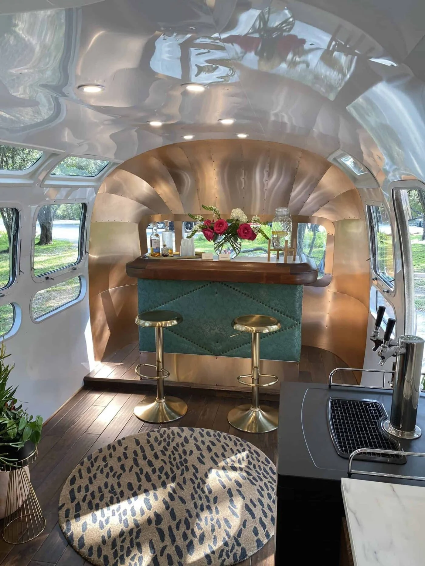 Interior of a converted camper with a bar area featuring a floral arrangement, two gold bar stools, a faucet, a sink, and a leopard print rug on wooden flooring. Windows provide natural light and a view of trees outside.
