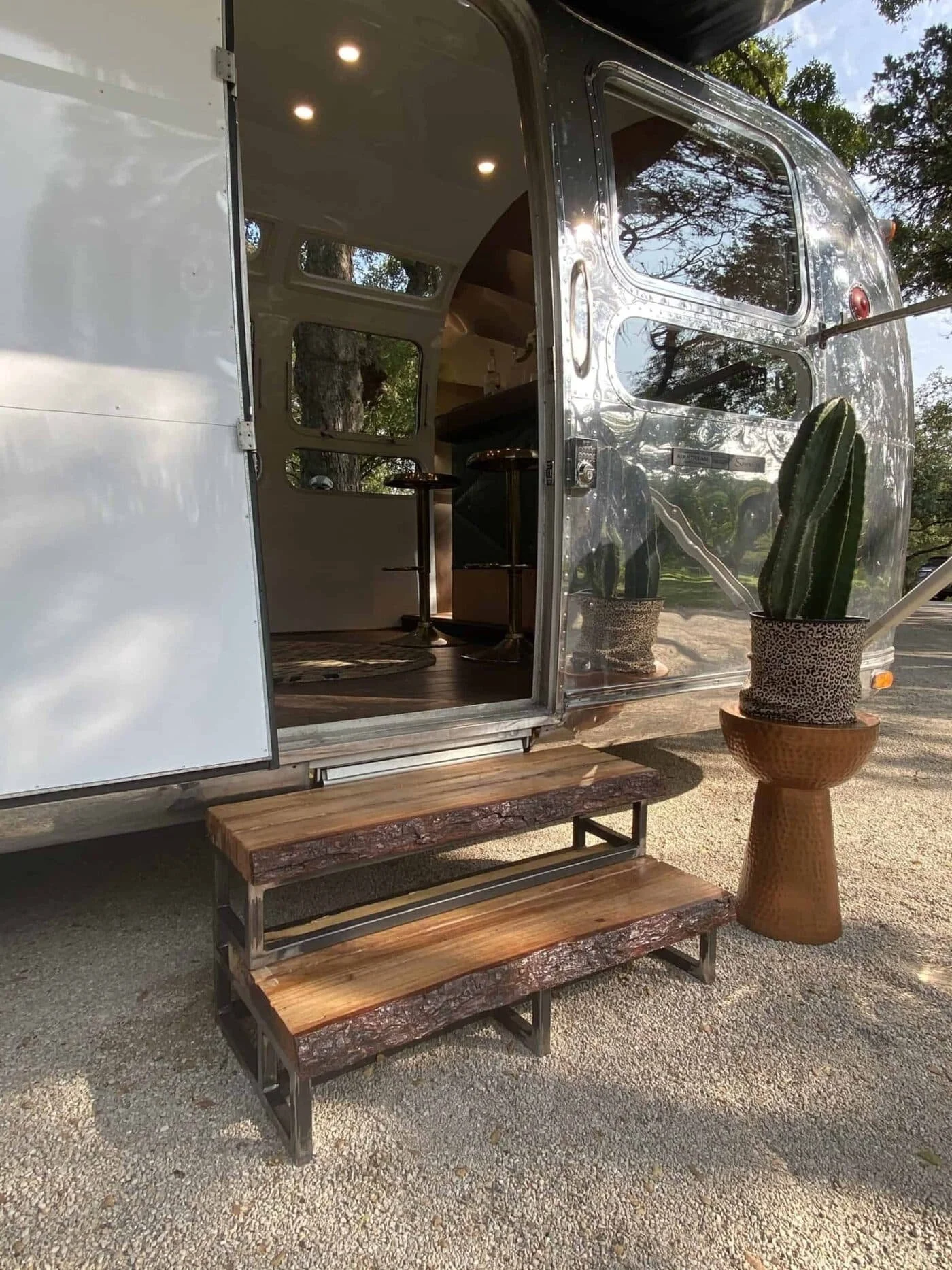 An Airstream travel trailer with open door, showing interior with seating and small table. Outside, a cactus in a pot is on a wooden stand, and a small wooden bench is on the gravel ground.