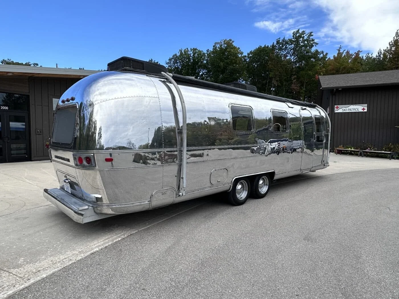 A shiny, vintage silver Airstream camper trailer parked on the street near a building with a ski patrol sign. The trailer reflects the sky, trees, and surrounding area.