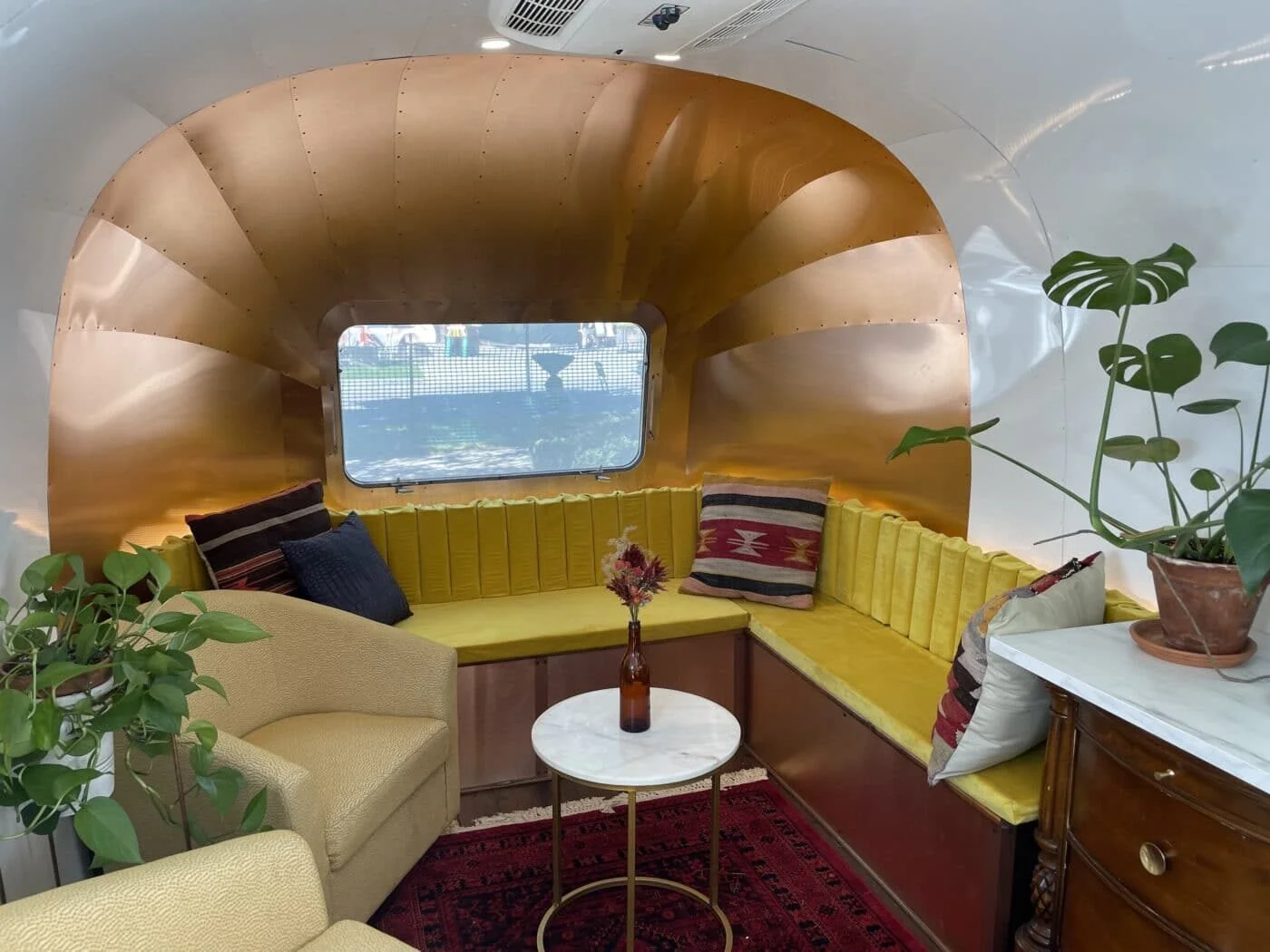 Interior of a retrofuturistic space with curved wooden wall, yellow cushioned seating, a small round marble table with a vase, an upholstered armchair, green plants, and a window showing water outside.