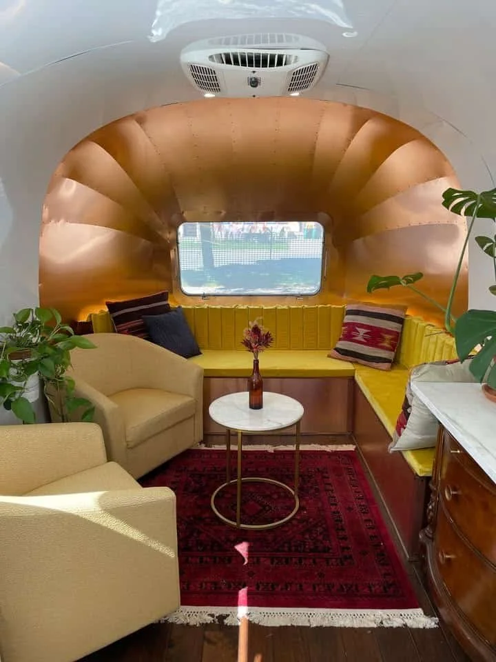 Cozy corner with yellow cushioned window seat and pillows, beige armchairs, a small round marble-top table with a flower vase, green plants, and a red patterned rug.