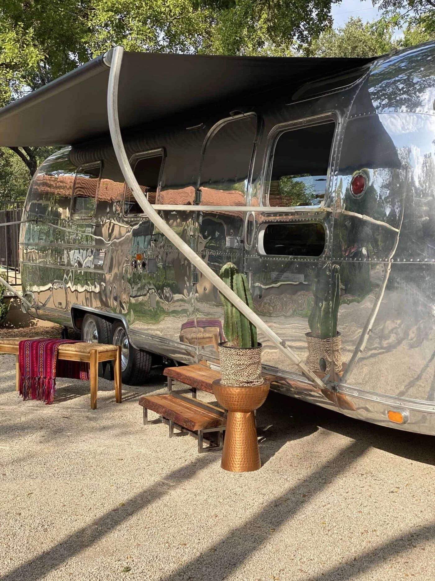 A shiny silver camper trailer with a large black awning extended outside. There are some small windows, and a cactus plant in top of a textured tall pot near the trailer. There is also a small wooden bench with a colorful blanket draped over it, and 